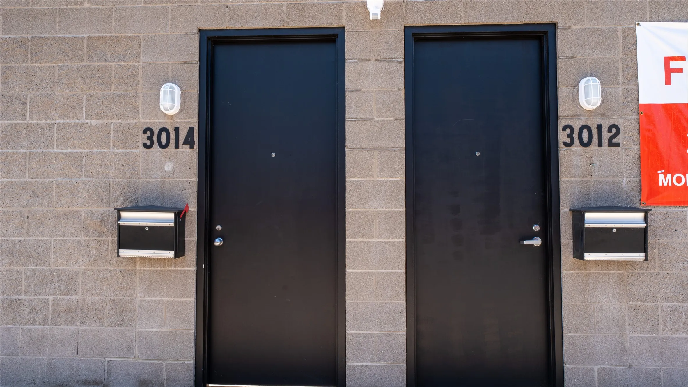 Exterior entryway featuring concrete masonry unit siding, matte black flush-mount entry doors, wall-mounted metallic mailboxes, and oval-shaped bulkhead lighting fixtures