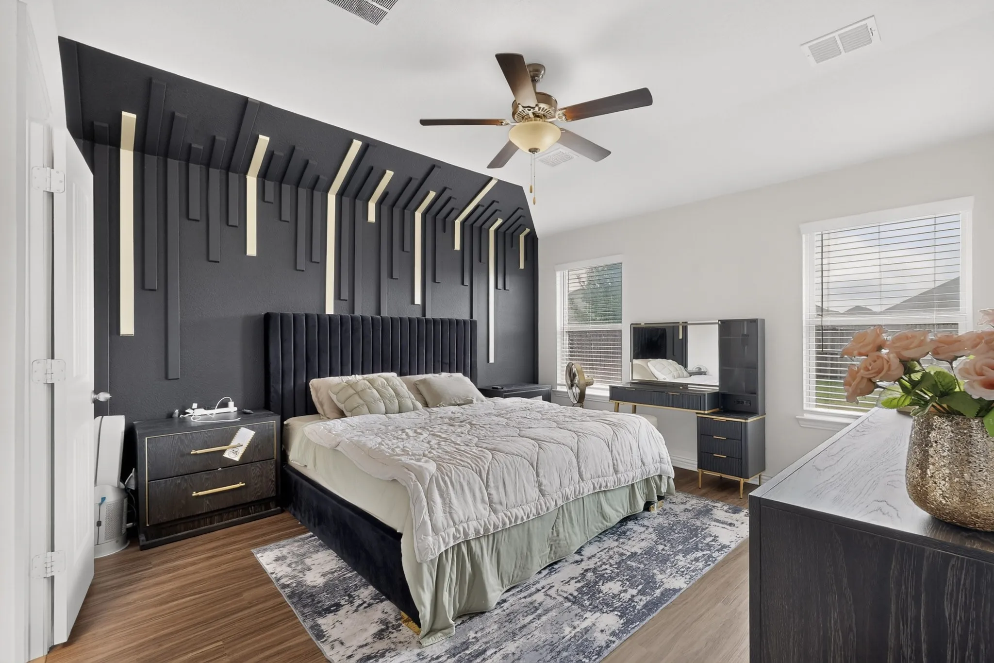 Custom dark-toned accent wall with vertical geometric wood molding, wood-finish flooring, ceiling fan with integrated lighting, and multiple windows with horizontal blinds