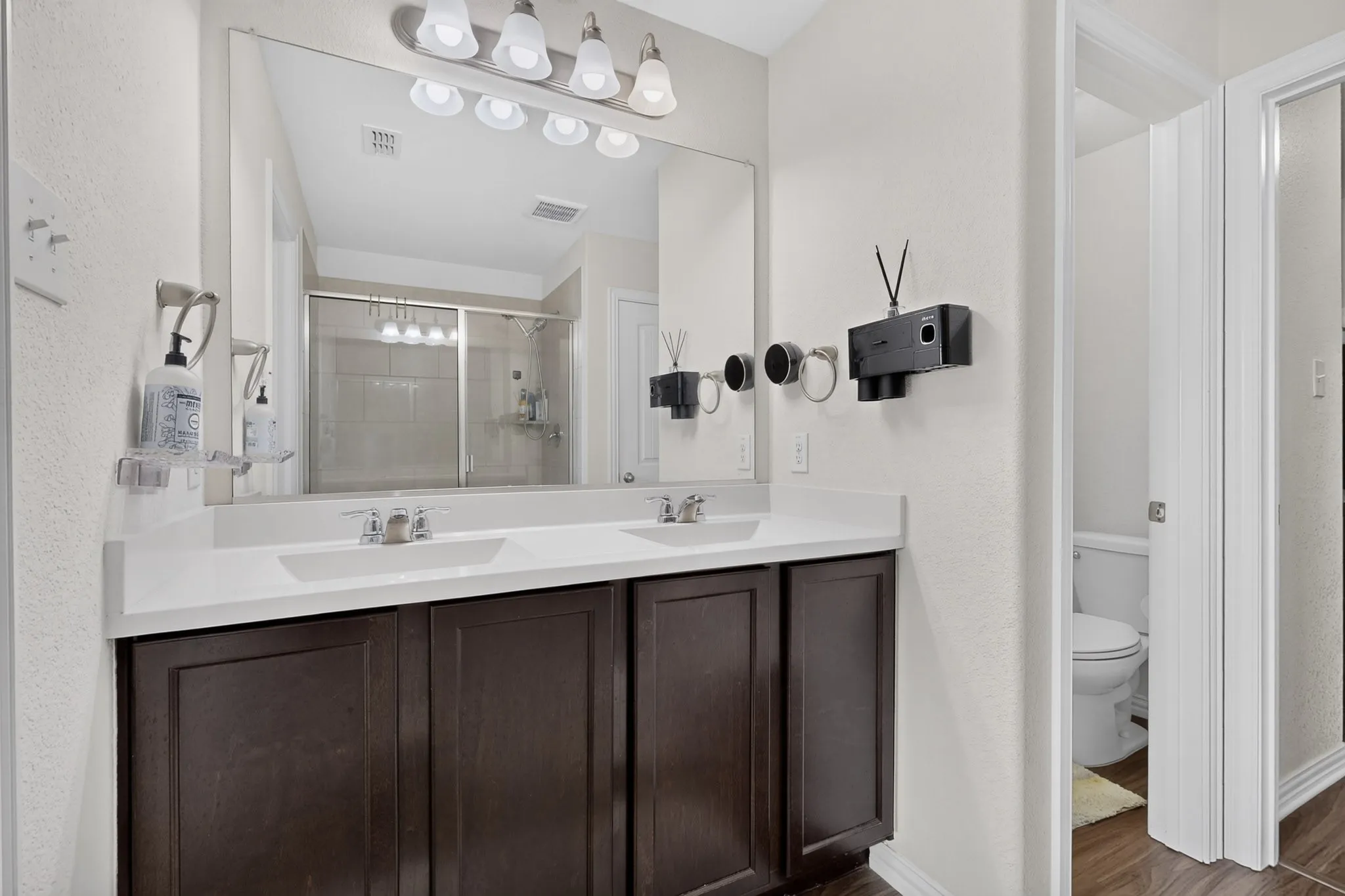 Dual-sink vanity featuring dark-finish cabinetry, light-toned countertops, and a full-width wall mirror with integrated multi-bulb lighting