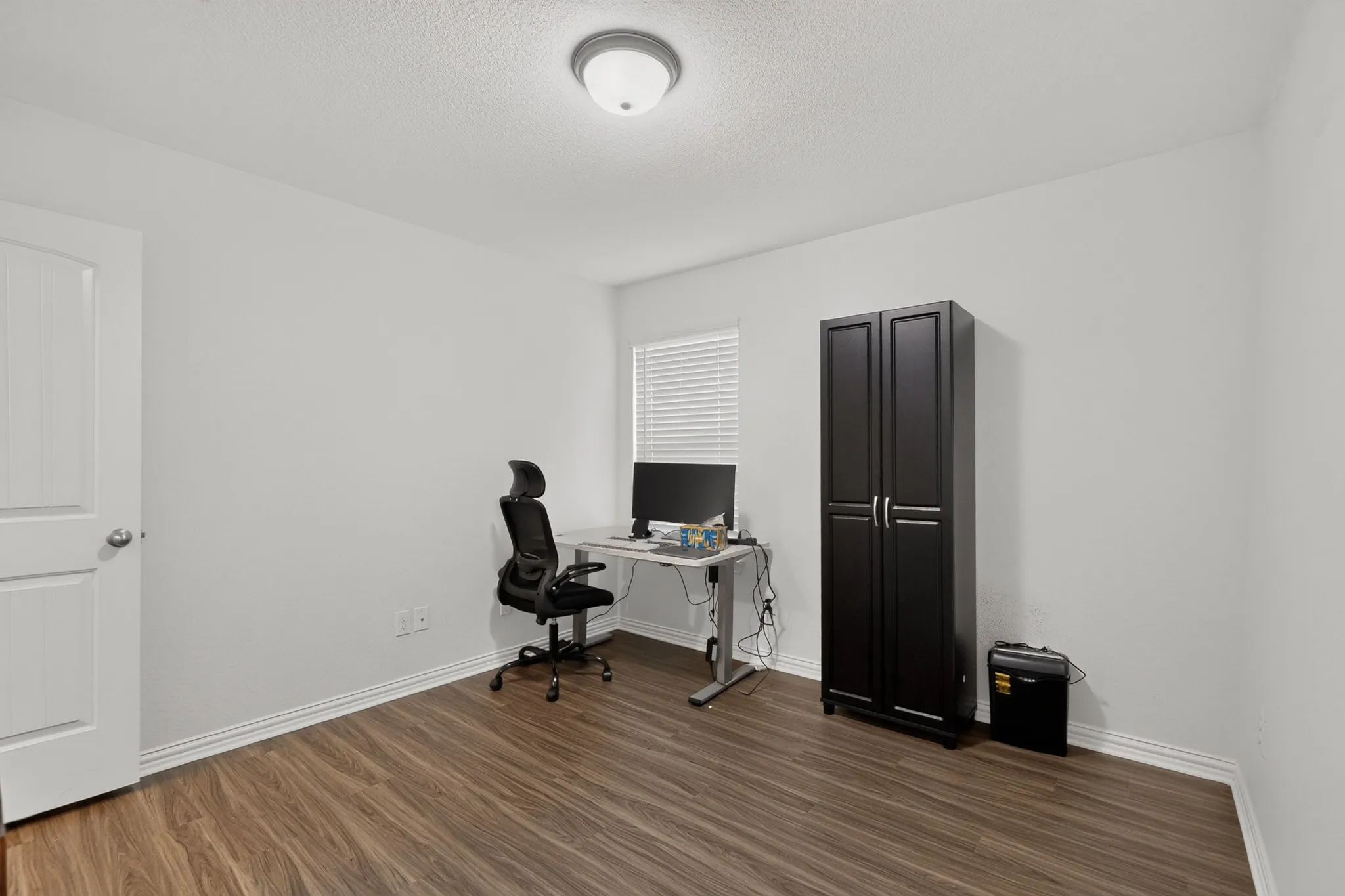 Room featuring wood-finish flooring, white walls, flush-mount ceiling lighting, and a window with horizontal blinds