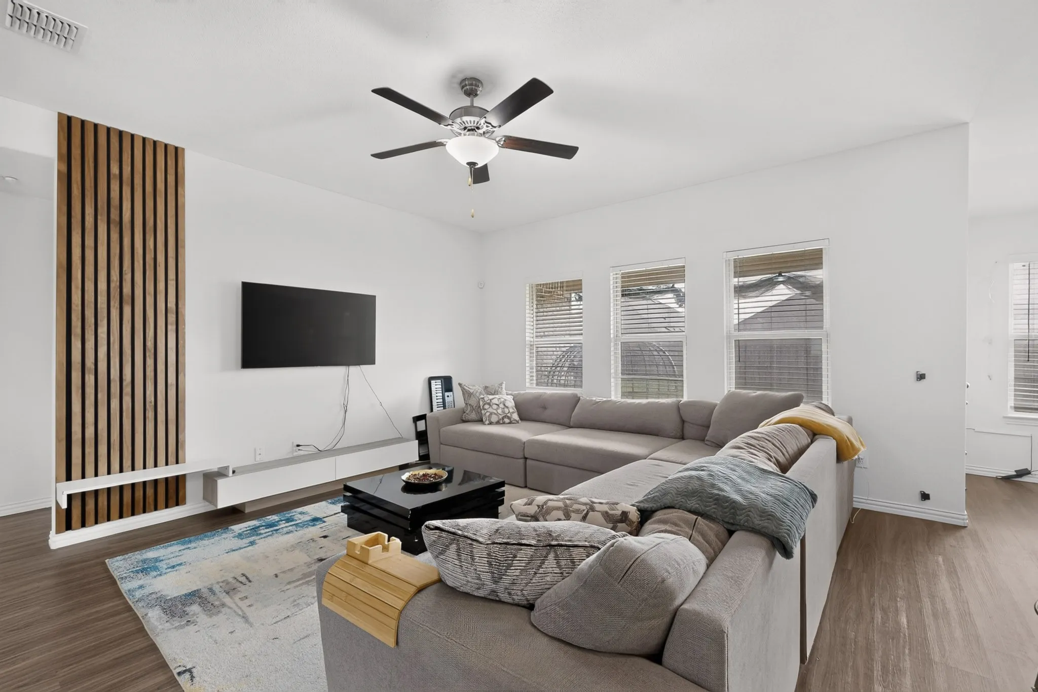 Living area featuring wood-finish flooring, a vertical wood slat accent wall, a floating media console, and a multi-blade ceiling fan with integrated light fixture