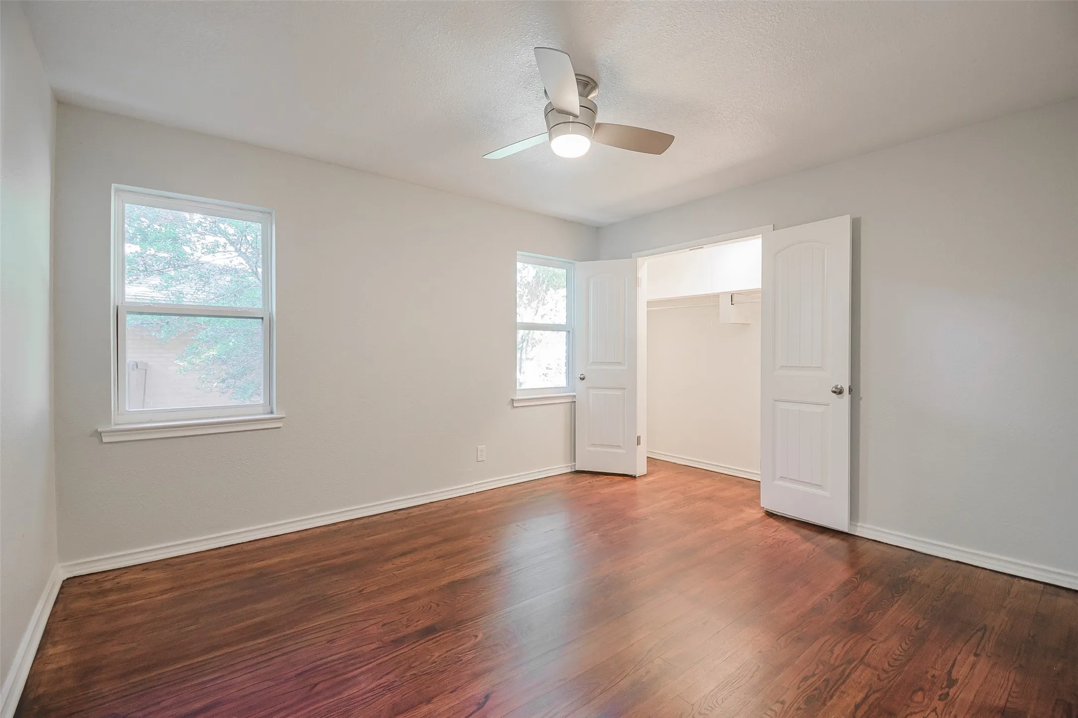 Room featuring wood-finish flooring, two double-hung windows, a ceiling-mounted light fixture with fan, and a double-door closet