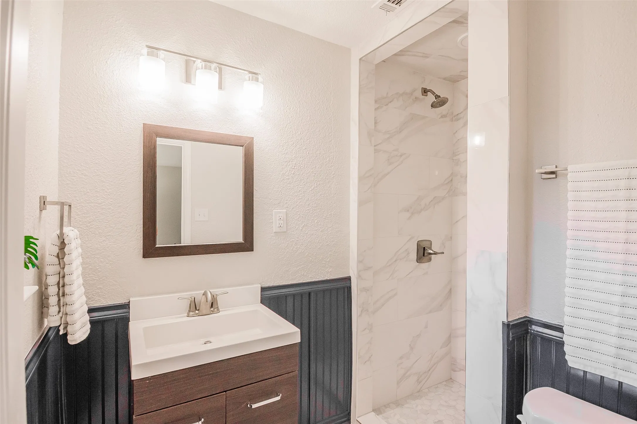 Bathroom featuring a vanity with a white rectangular sink, dark wood-finish cabinetry, wainscot paneling, a shower enclosure with stone-patterned tile, and a three-light wall-mounted fixture