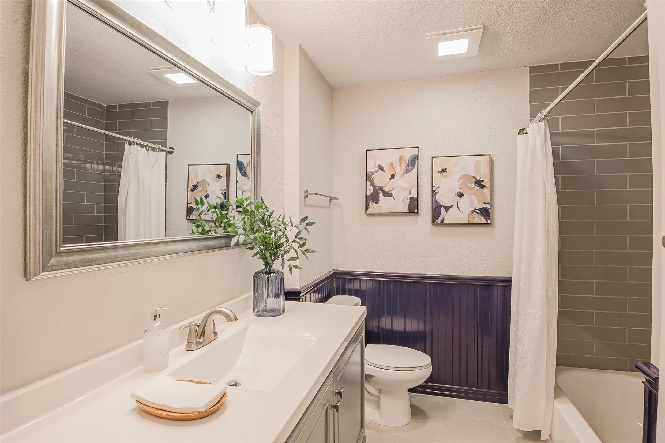 Bathroom featuring a vanity with an integrated basin, a large framed mirror, dark wood-tone wainscoting, and a tiled shower surround