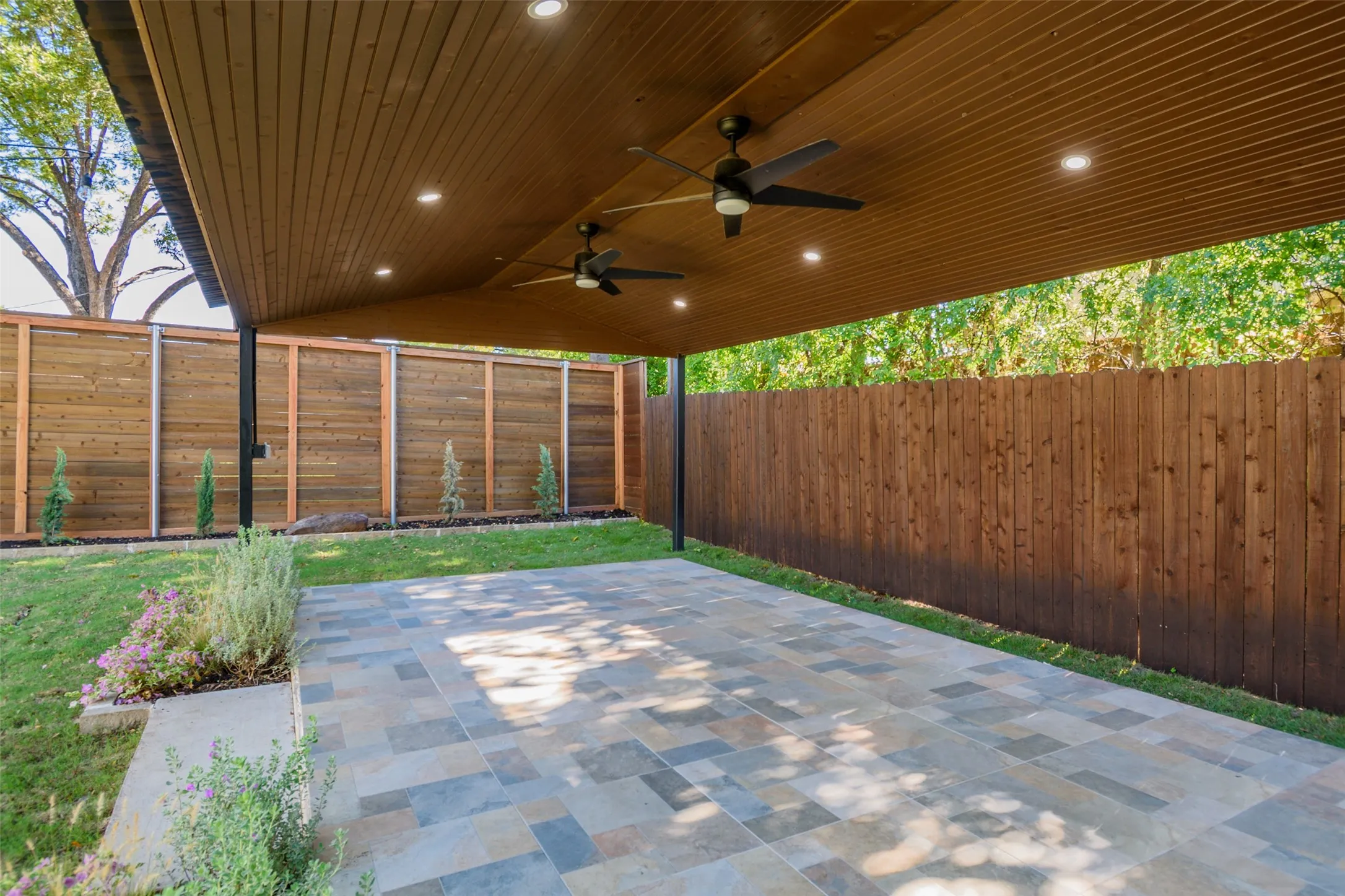 Covered patio featuring a wood-plank ceiling, recessed lighting, dual ceiling fans, and multi-tonal stone flooring, enclosed by a wood privacy fence