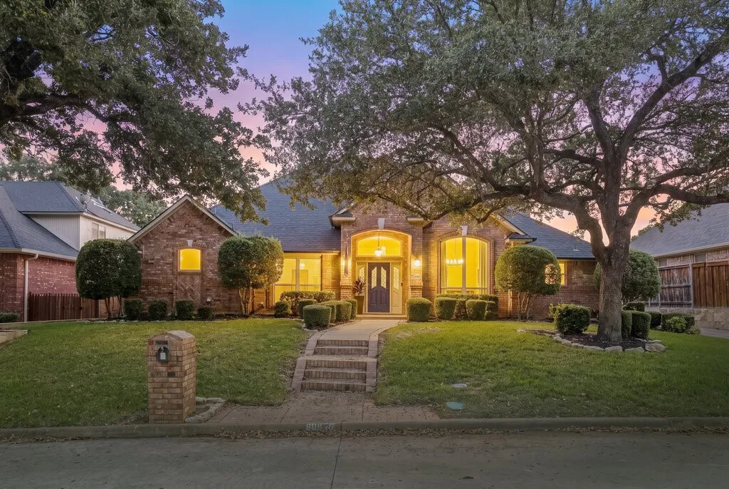 Brick exterior featuring an arched entryway, gabled roofline, manicured landscaping, and mature canopy trees