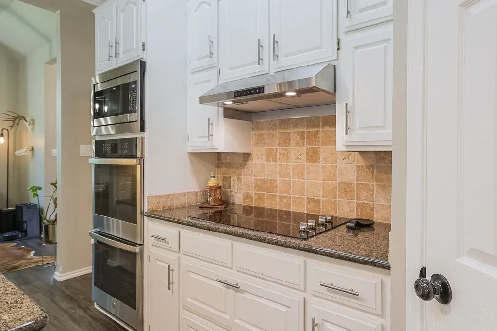 Kitchen featuring white cabinetry, stainless steel double wall ovens, glass cooktop, stone-finish tile backsplash, and dark stone-surface countertops