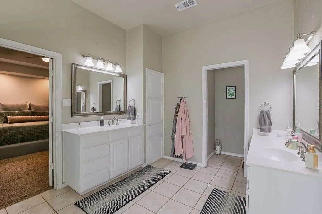Dual vanities featuring white cabinetry, solid surface countertops, rectangular under-mount sinks, chrome hardware, and wall-mounted vanity lighting