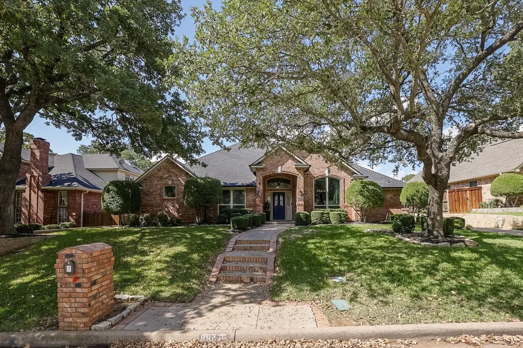 Brick exterior residence featuring a front gabled roofline, arched entryway, masonry mailbox, and tiered brick walkway set within a landscaped lawn shaded by mature trees