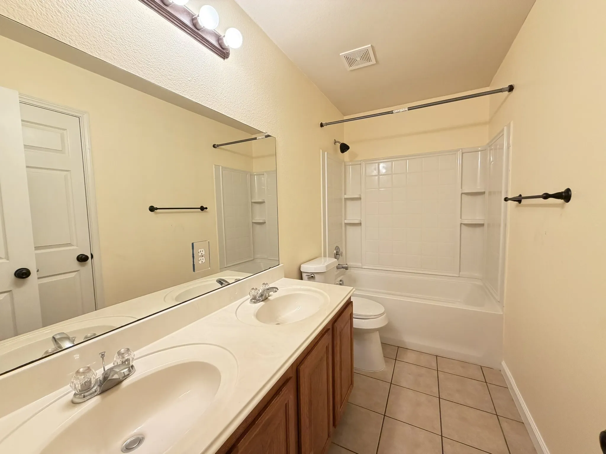 Dual-basin vanity featuring an integrated countertop, wood cabinetry, and a full-width wall mirror, complemented by a combination tub and shower with tile-patterned surround and ceramic floor tiling
