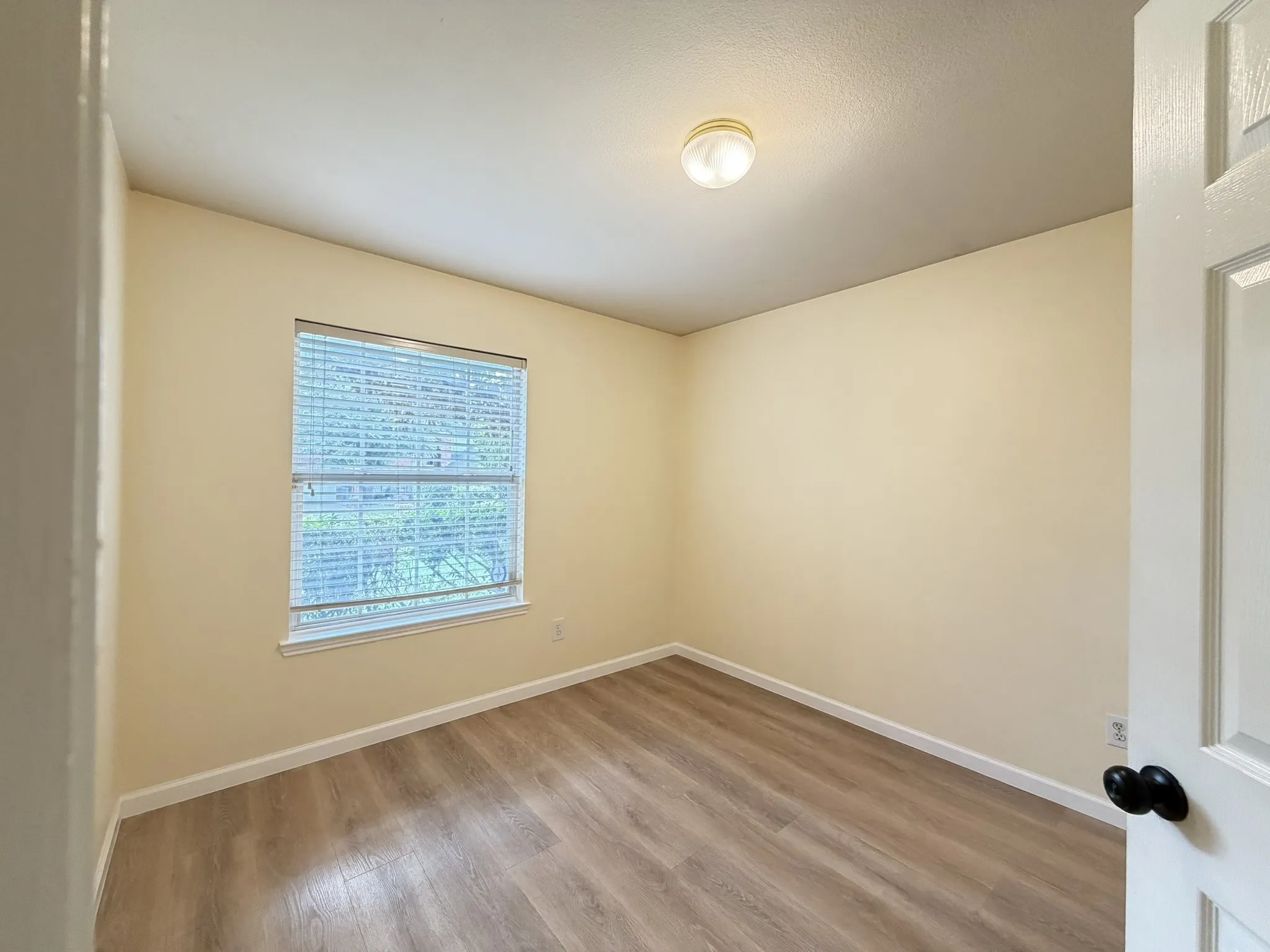 Neutral-toned interior space featuring wood-finish flooring, a single window with horizontal blinds, and a circular ceiling-mounted light fixture