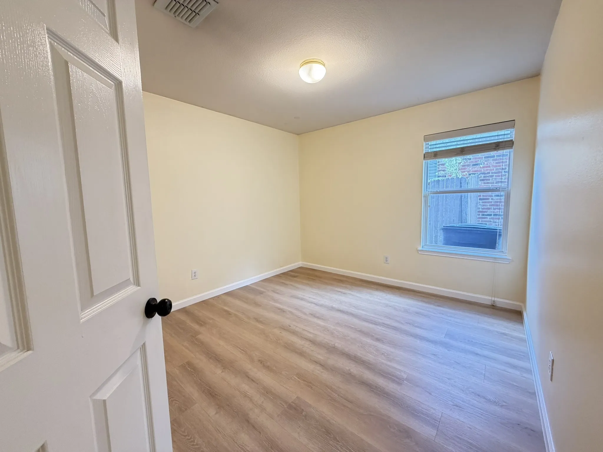 Room featuring neutral wall paint, wood-finish flooring, a single-hung window with a roller shade, and a central flush-mount light fixture
