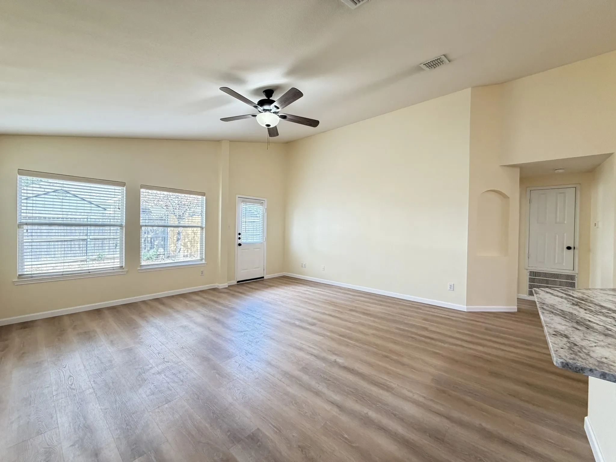 Vaulted ceiling with an attached light fixture and ceiling fan, wood-finish flooring, two large windows with horizontal blinds, and a built-in recessed arched wall niche