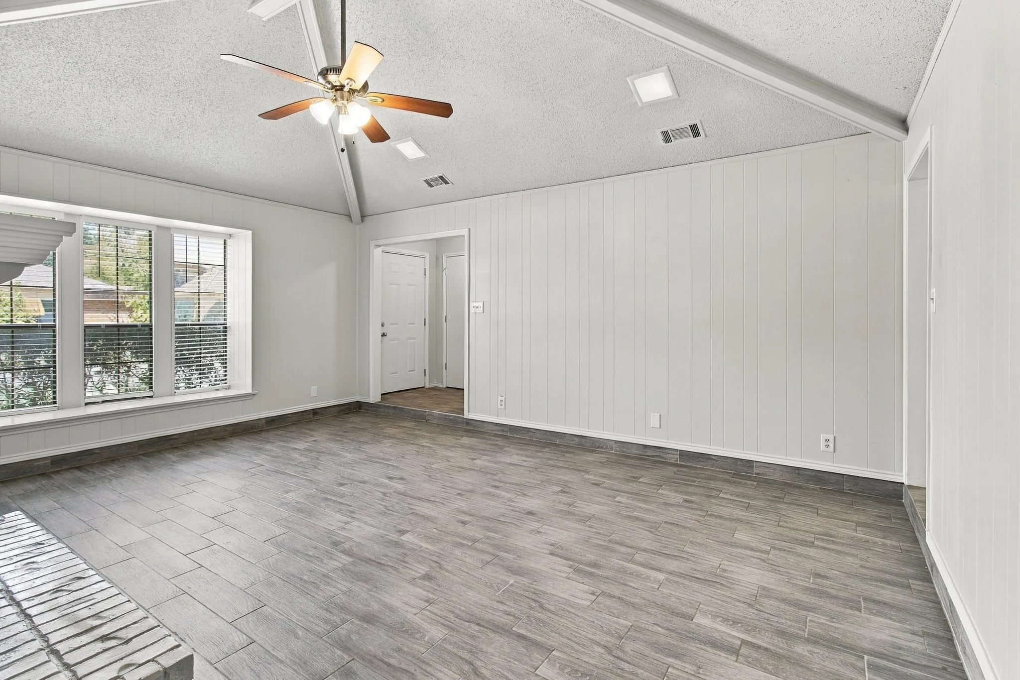 Vaulted ceiling featuring a central wood-finish ceiling fan, vertical wall paneling, wood-finish flooring, and multiple window panes