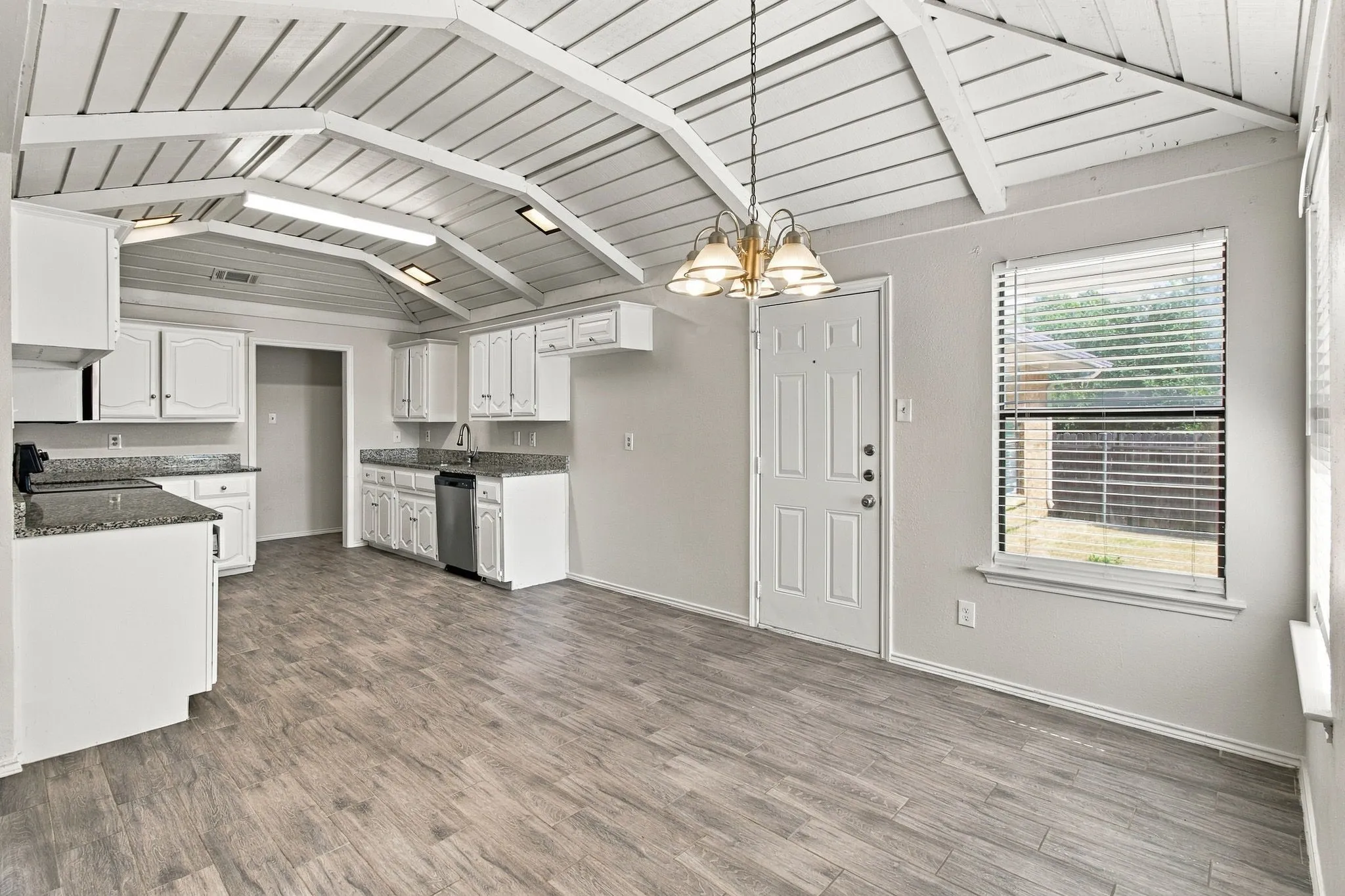 Vaulted ceiling with exposed white beams, wood-finish flooring, white cabinetry, stone-finish countertops, and an integrated multi-bulb chandelier