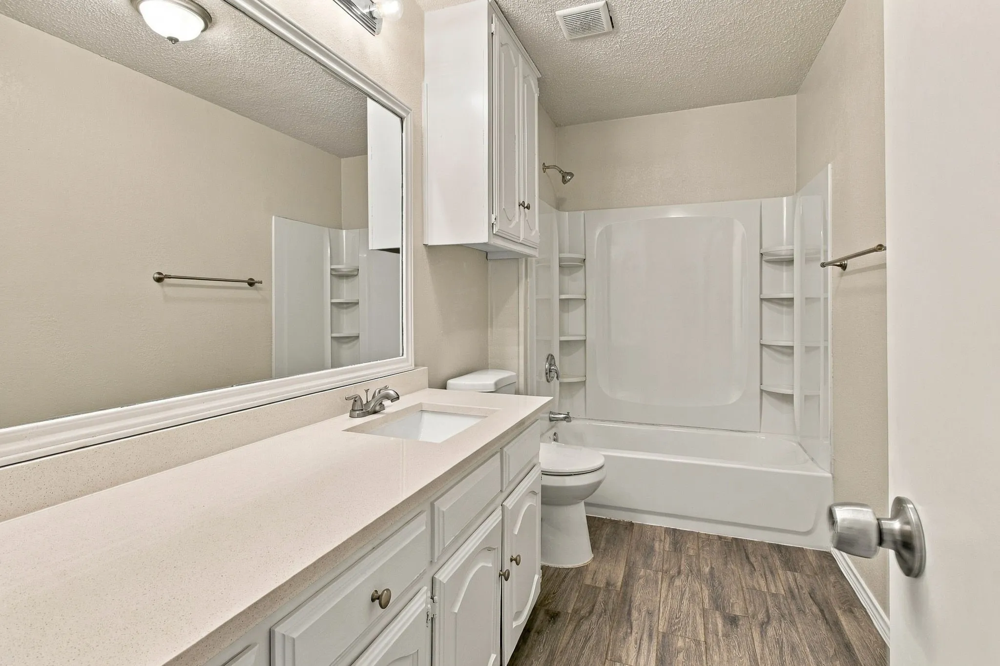 Bathroom featuring a single-sink vanity with a solid-surface countertop, full-width mirror, wood-finish flooring, and a tub-shower combination with integrated shelving