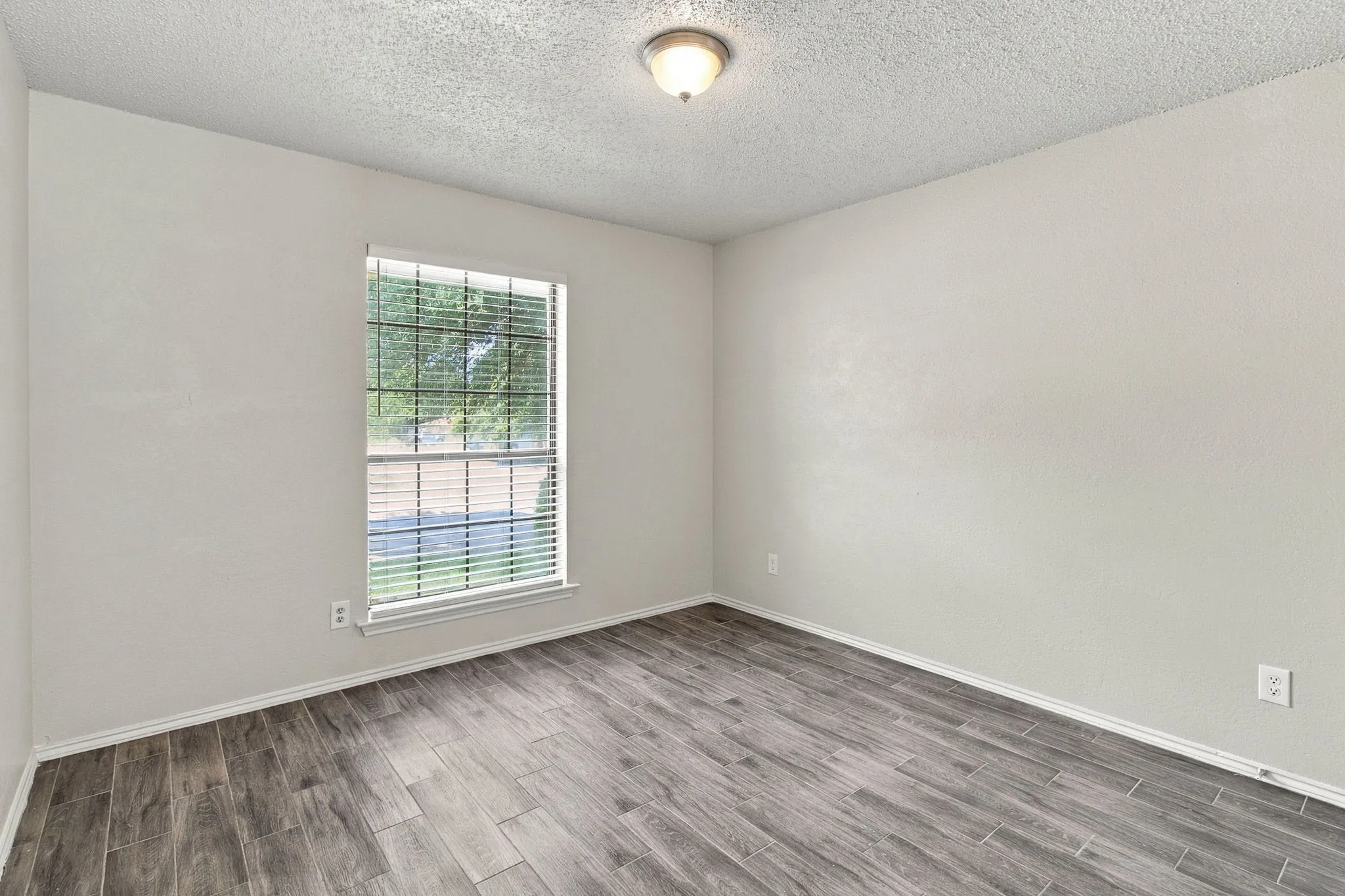 Neutral-toned walls, wood-finish tile flooring, a large window with horizontal blinds, and a central flush-mount ceiling light fixture