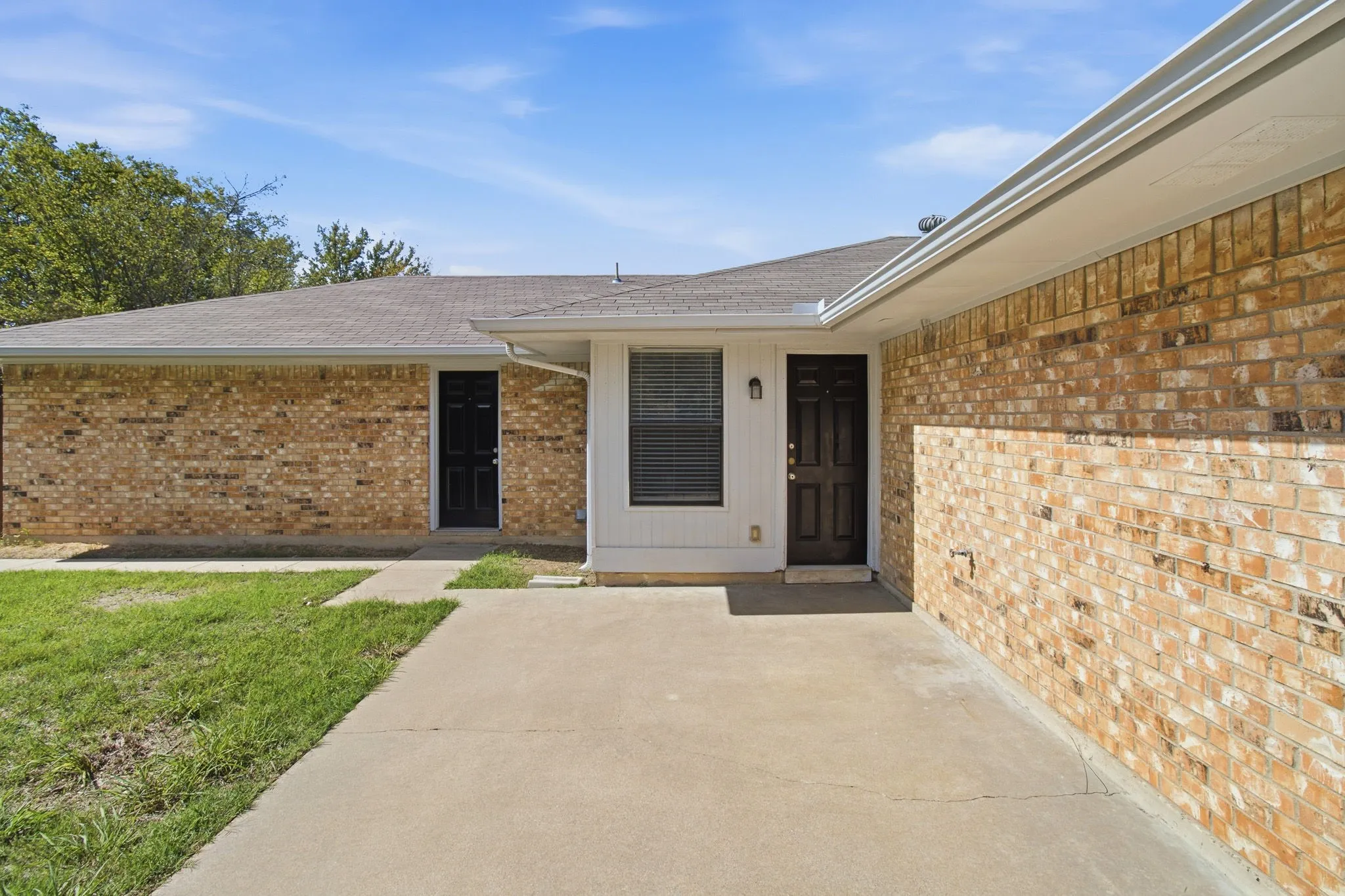 Single-story exterior featuring multi-toned brick siding, a neutral-toned shingle roof, two solid-panel entry doors, and a concrete patio