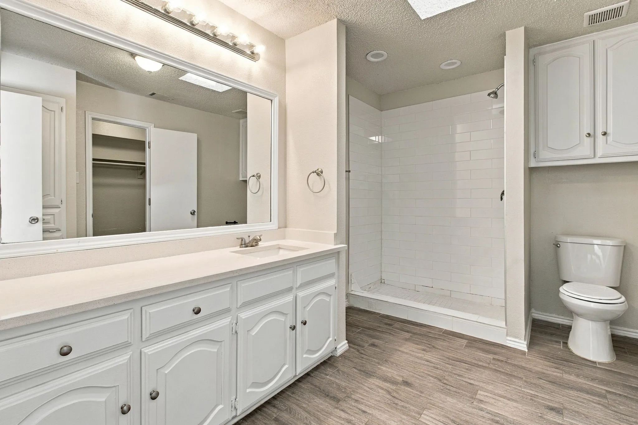 Bathroom featuring a vanity with a solid-surface countertop, white cabinetry, subway-tiled walk-in shower, wood-finish flooring, and wall-mounted cabinetry