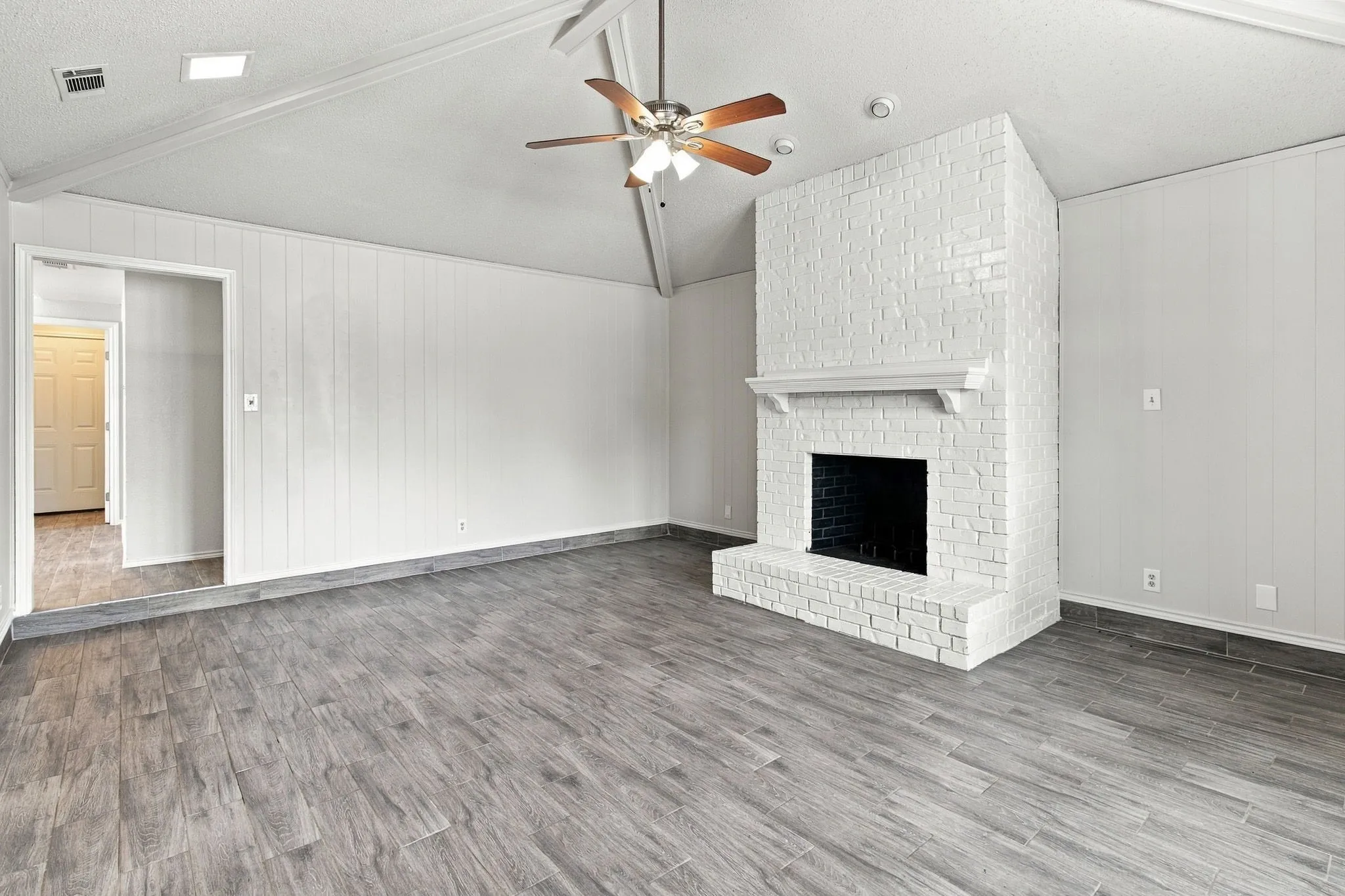 Vaulted ceiling with an integrated ceiling fan, white-painted brick fireplace with a decorative mantel, and wood-finish flooring