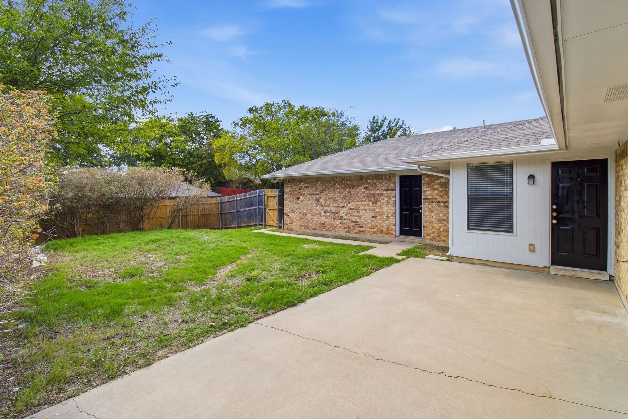 Rear exterior featuring a concrete patio, brick and siding facade, two dark-toned entry doors, and a fenced yard with mature landscaping