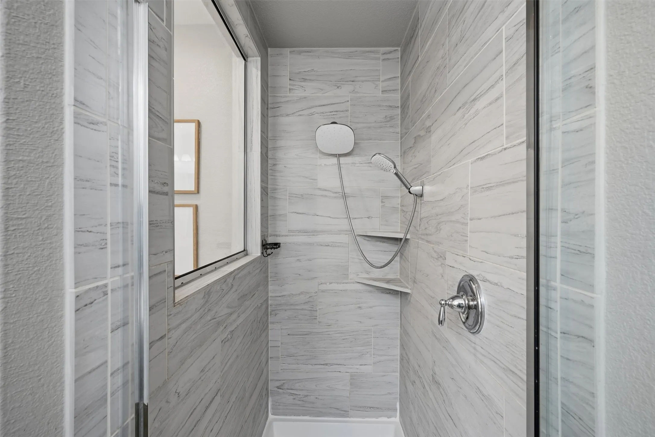 Detailed view of the primary bathroom shower featuring floor-to-ceiling marble-look tile, built-in corner shelves, and dual shower heads including a handheld wand. Glass enclosure maintains bright sightlines to the rest of the bath.