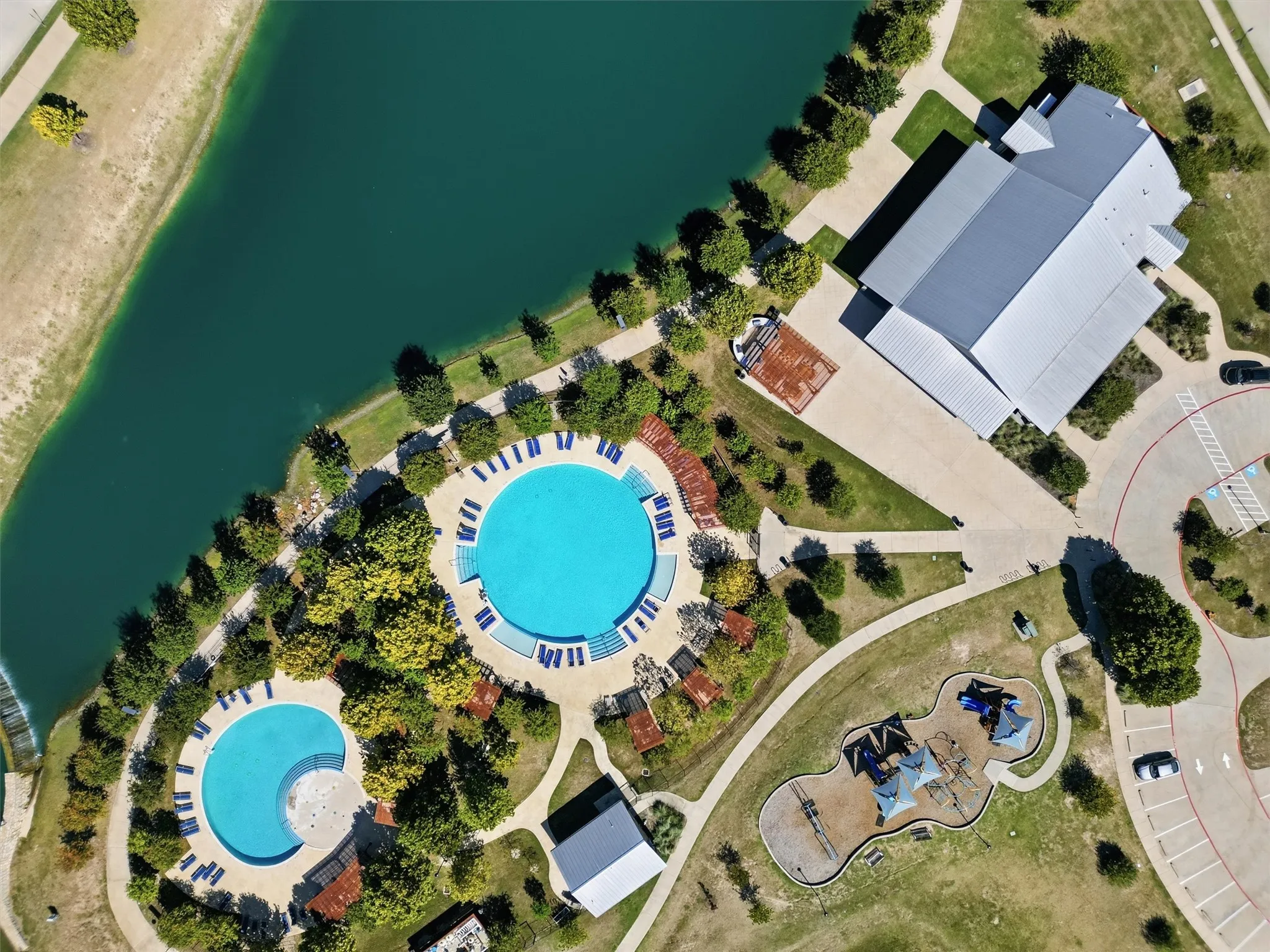 Direct top-down aerial view detailing the resort-style amenities layout. Showcases the side-by-side dual circular swimming pools surrounded by sun decks, shaded pergolas, connected walking paths, playground zone, and the private event clubhouse along the scenic water's edge.