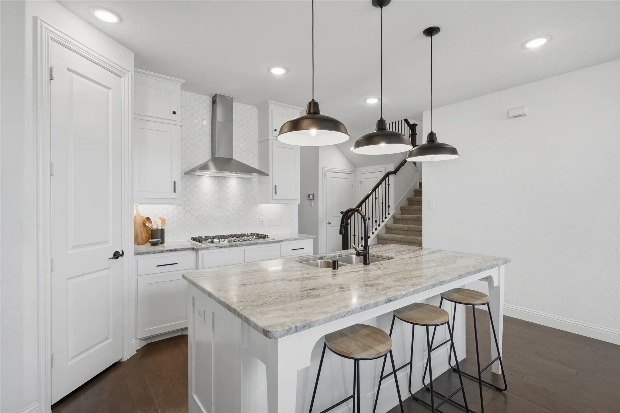 Great view of the kitchen island highlighting granite countertops, an undermount double sink, and pendant lighting. Shows the gas cooktop with stainless vent hood, geometric tile backsplash, and pantry door.