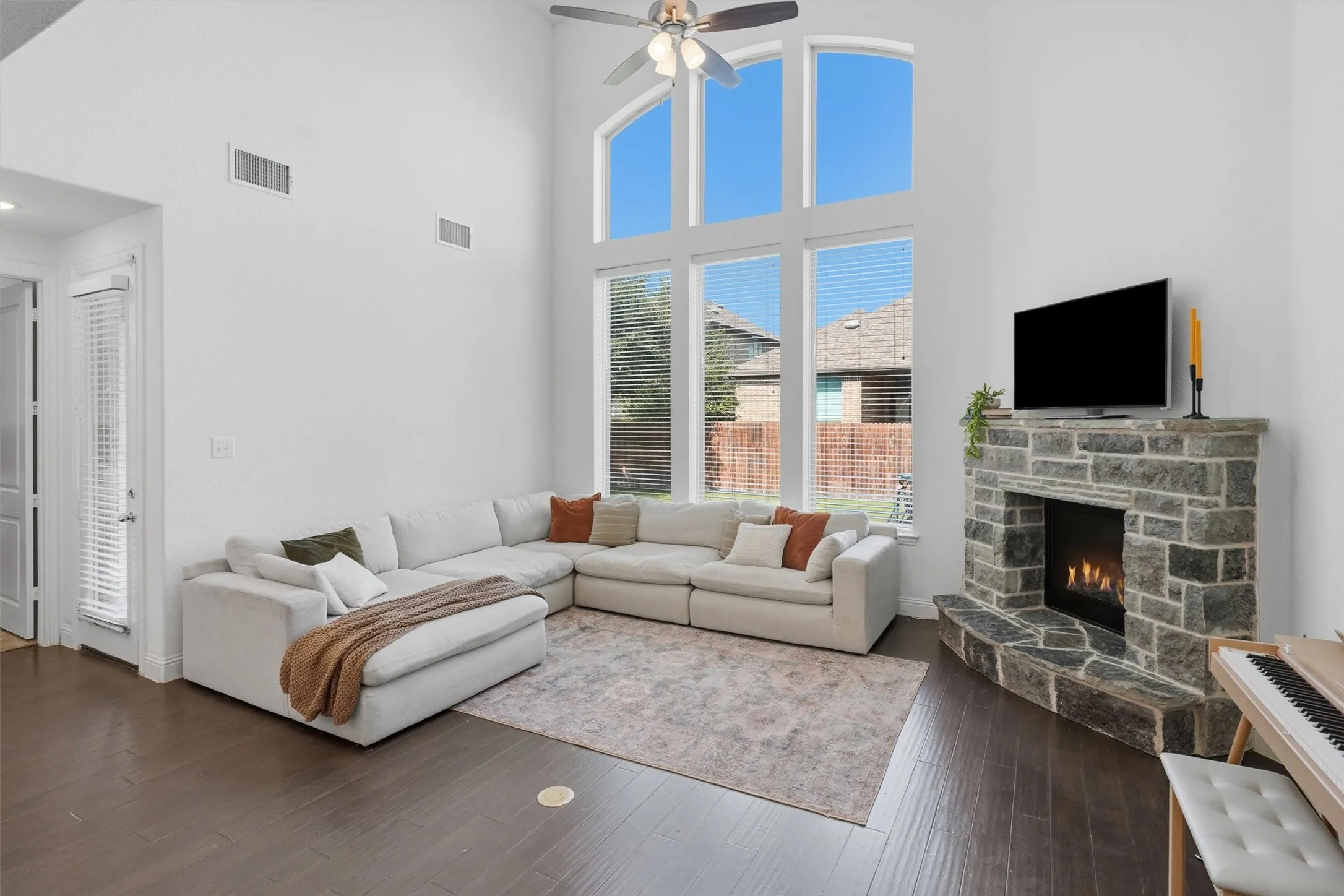 Dramatic two-story living room with soaring vaulted ceilings and floor-to-ceiling arched windows. Features a stacked-stone corner gas fireplace, dark wood flooring, and a ceiling fan.