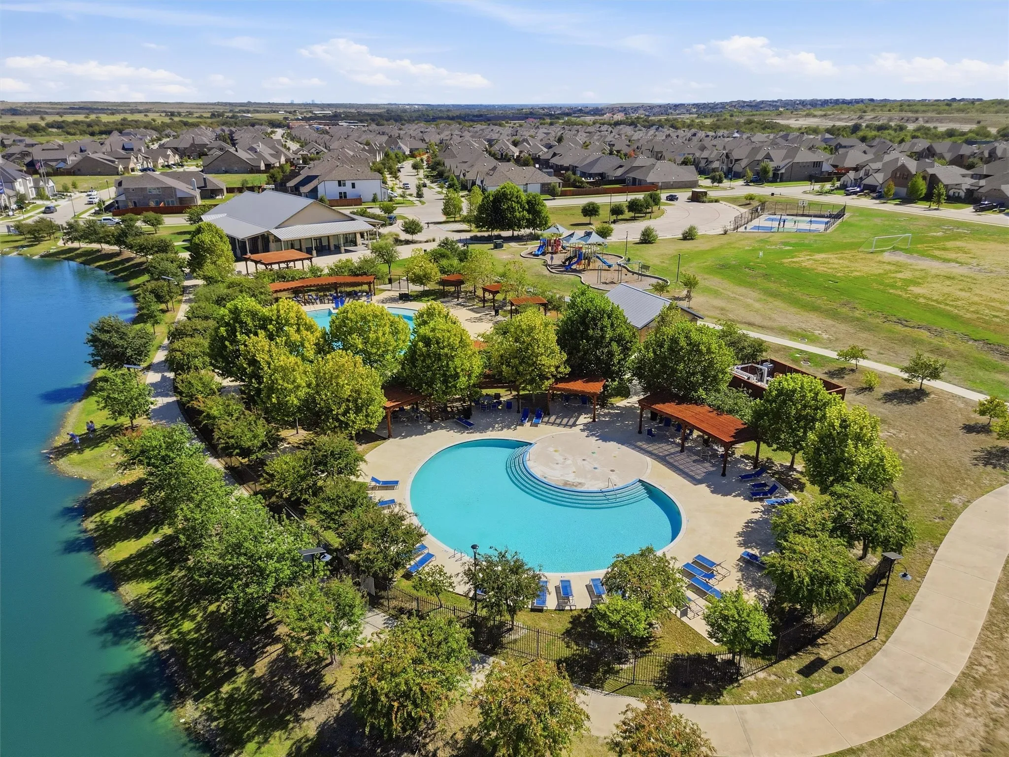 Stunning elevated aerial view highlighting the community's dual resort-style swimming pools situated along the edge of the scenic fishing pond. Displays the surrounding walking trails, shaded pergolas, play structures, sports courts, and private clubhouse.