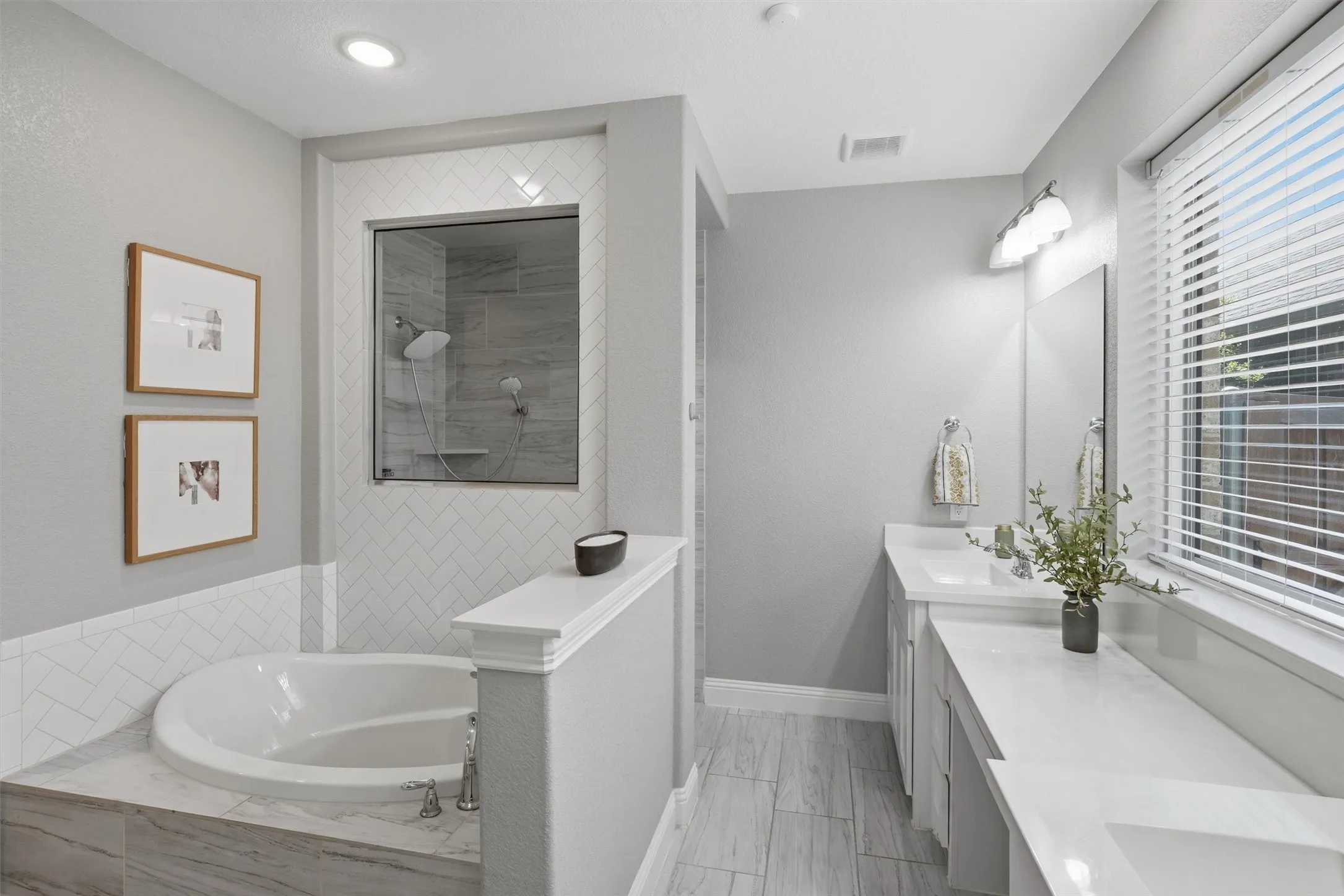Spa-inspired primary bathroom featuring a deep garden soaking tub, white herringbone wall tile, and a glass-enclosed walk-in shower. Includes dual vanities with a dedicated seated makeup area and a large window for light