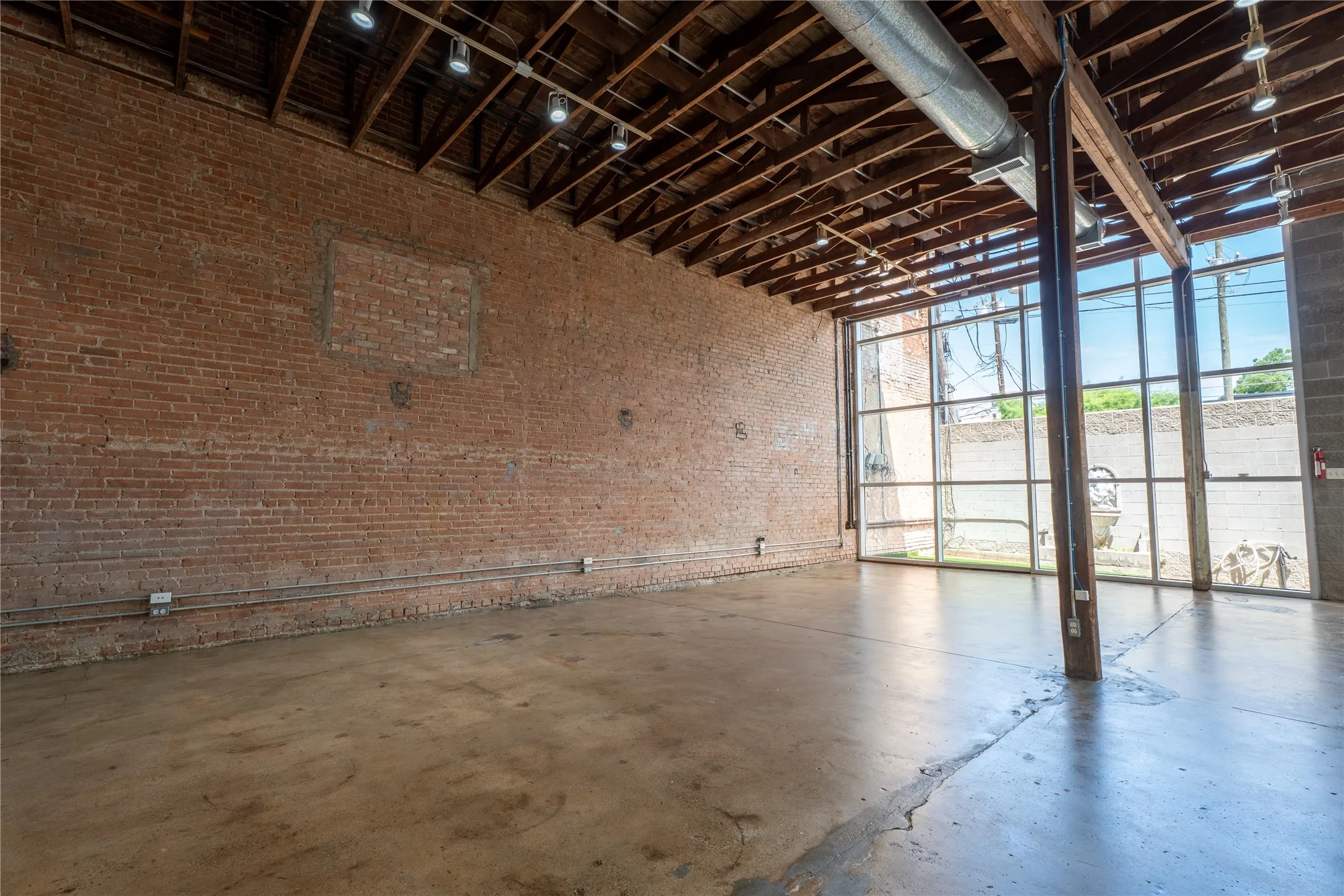 Industrial-style interior featuring exposed red brick walls, high ceilings with exposed structural beams, polished concrete flooring, full-height glass window wall, and industrial track lighting