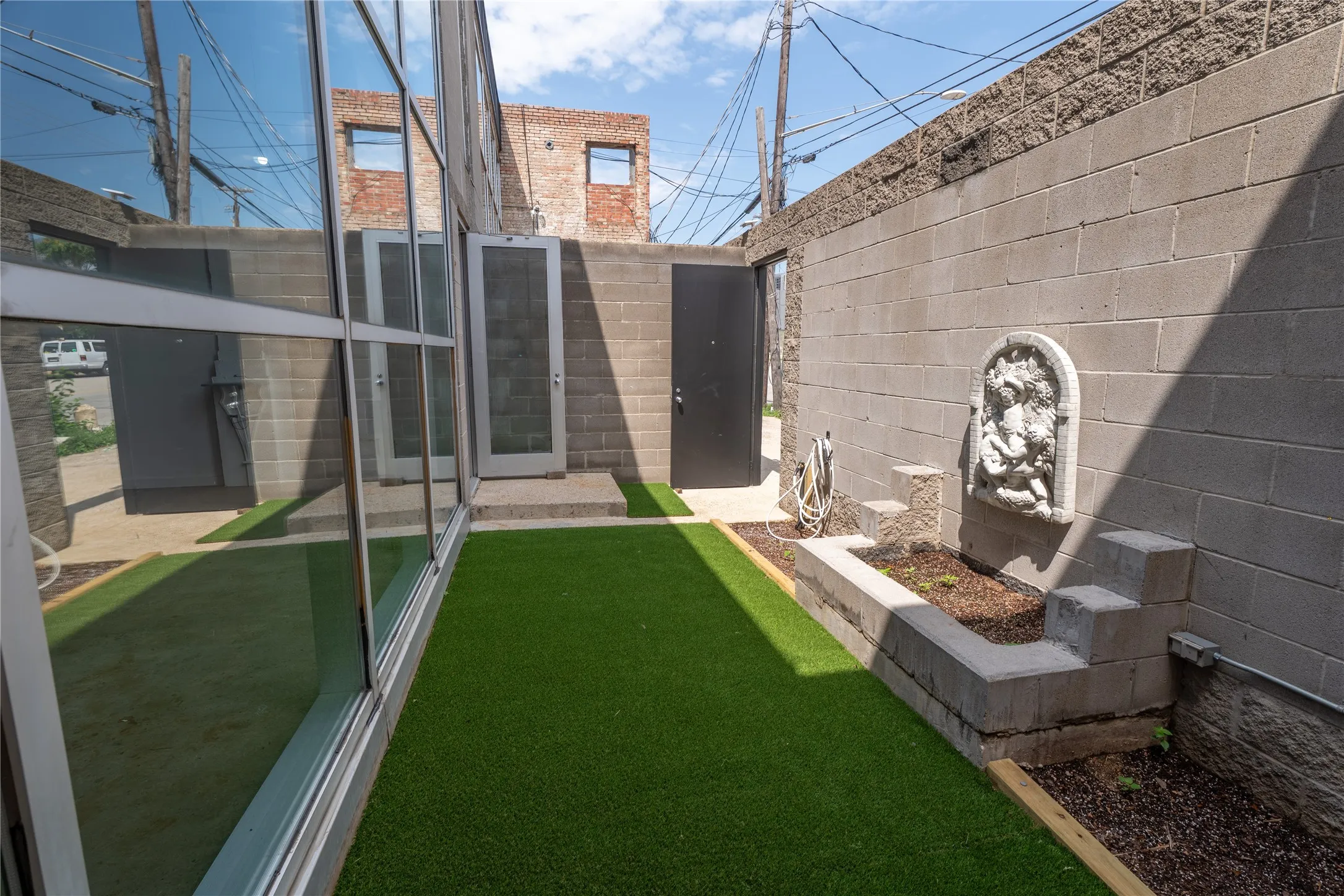 Enclosed courtyard featuring full-height glass window walling, concrete masonry unit privacy walls, integrated wall fountain with sculptural relief, and synthetic turf surfacing