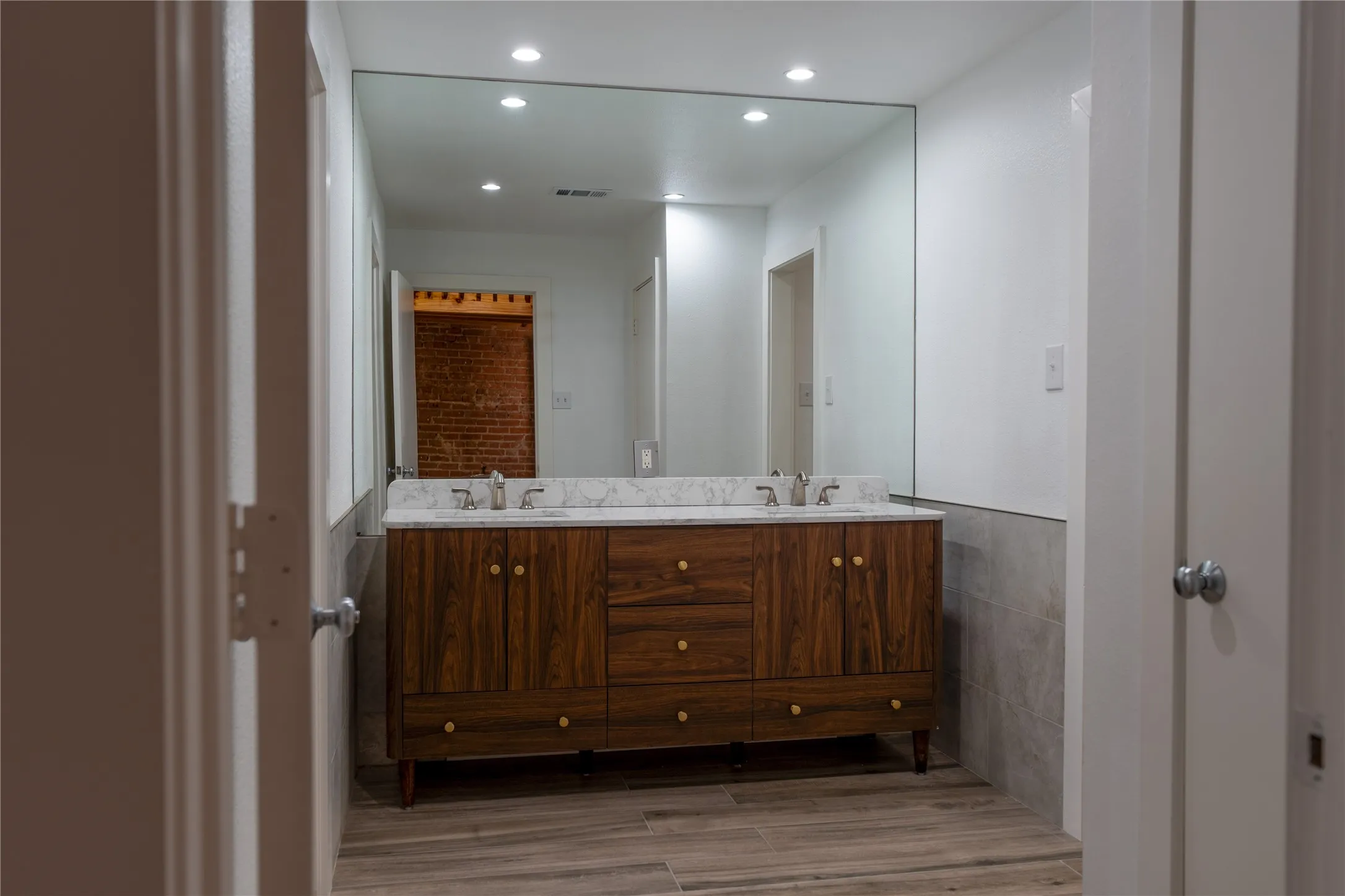 Dual-sink vanity with wood-finish cabinetry, stone countertop, integrated lighting, and full-width wall mirror over tile wainscoting