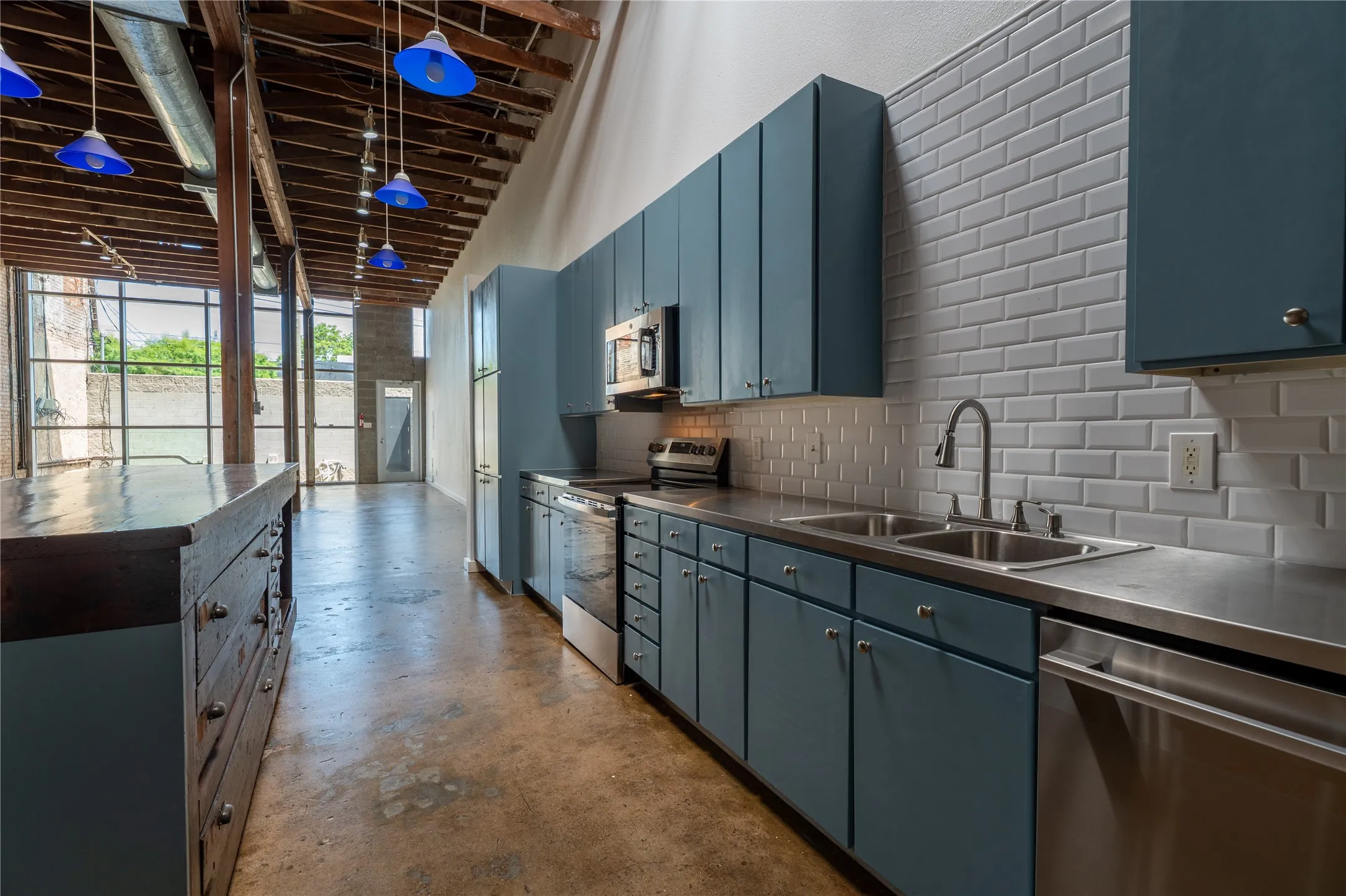Exposed timber ceiling beams, polished concrete flooring, stainless steel appliances, blue-toned cabinetry, and white subway tile backsplash