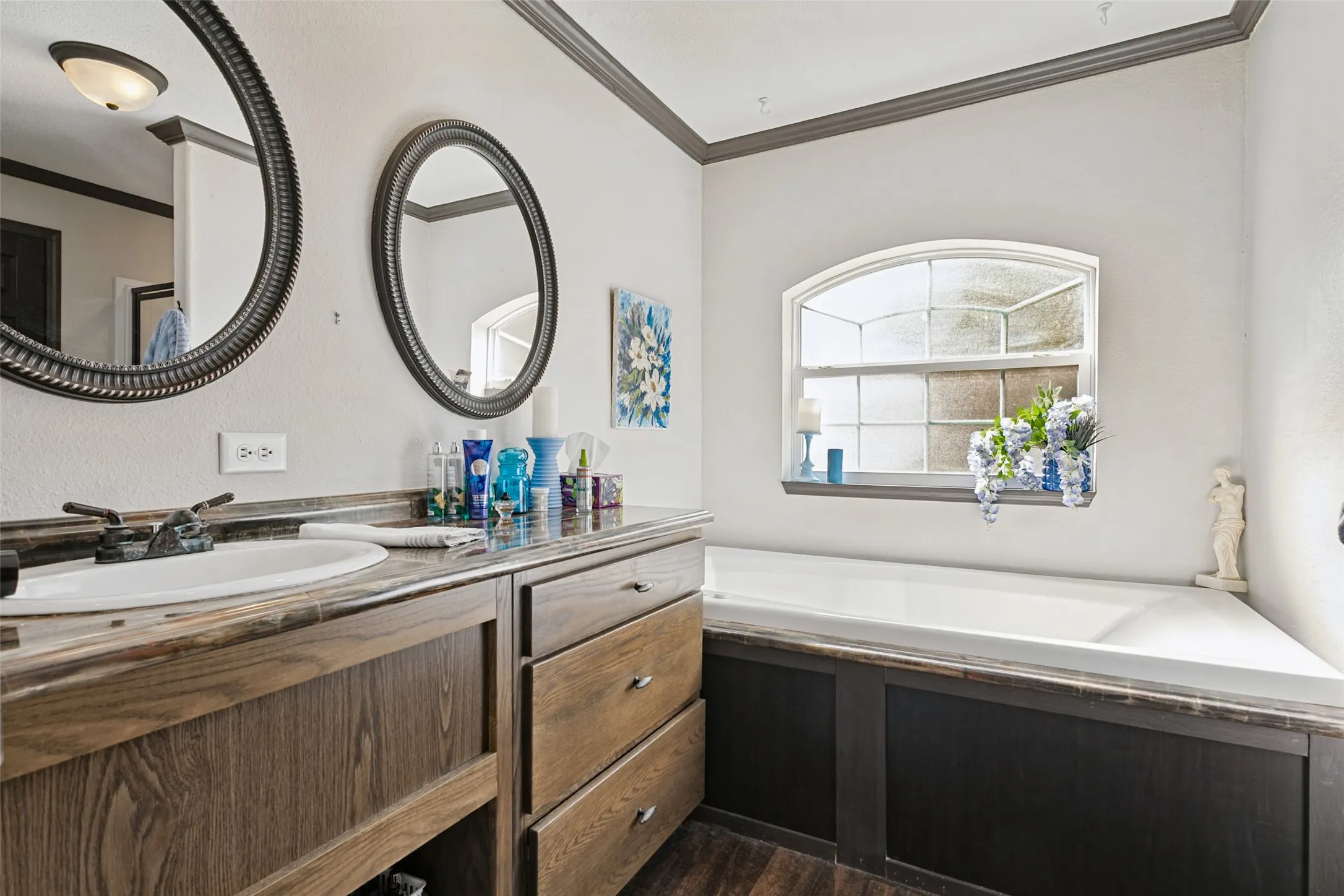 Wooden cabinetry vanity with a stone countertop, dual oval wall-mounted mirrors, and deep-soaking bathtub set beneath an arched window