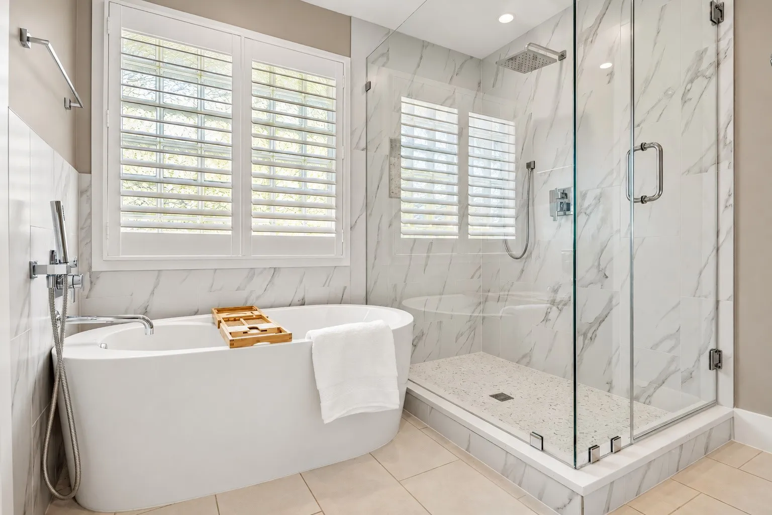 Freestanding soaking tub with chrome handheld faucet, frameless glass-enclosed shower, stone-finish wall tile, and custom window shutter