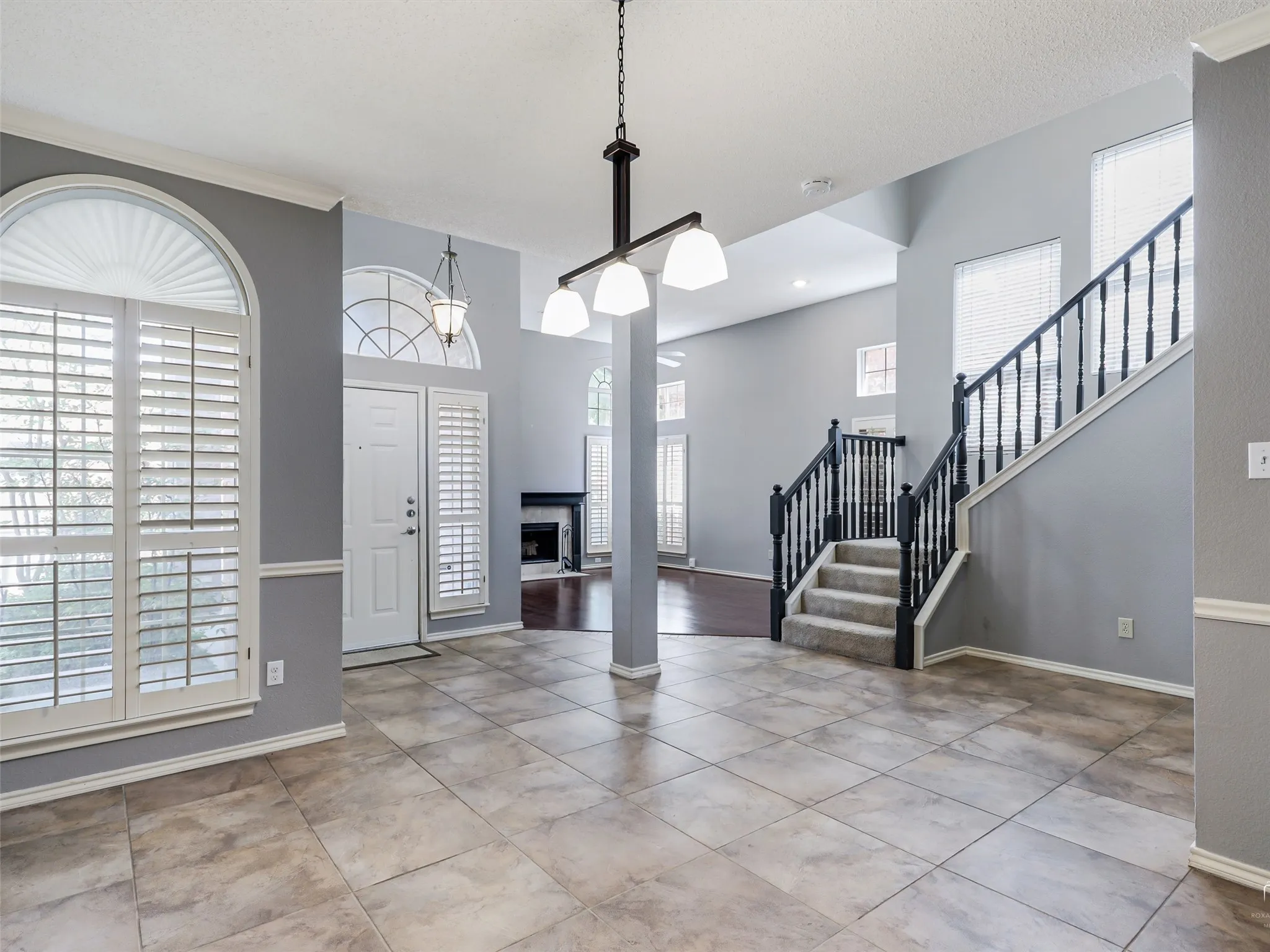 High-ceiling interior featuring tile flooring, arched windows with plantation shutters, a multi-light chandelier, and a staircase with a dark-finished handrail and balusters