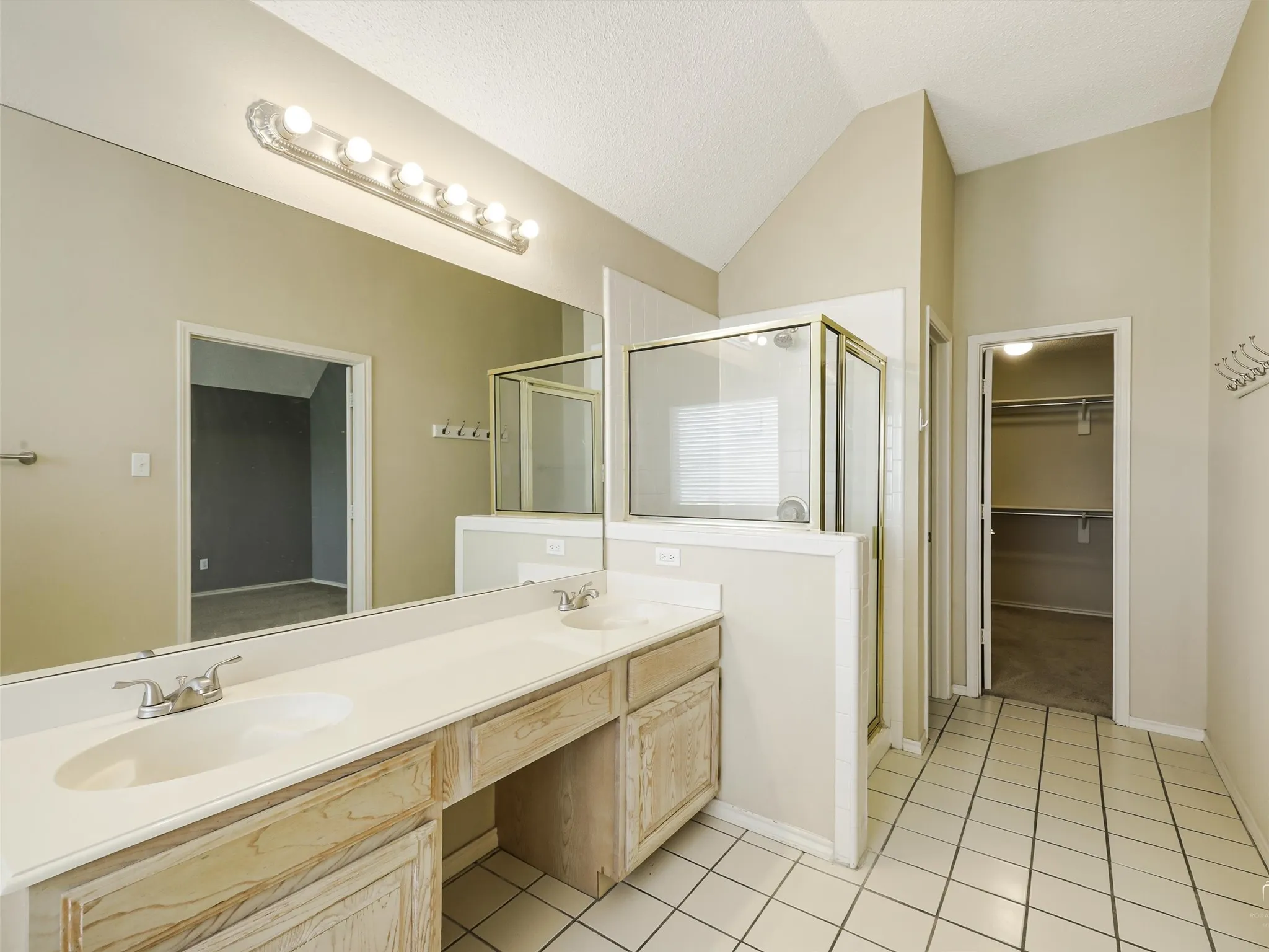 Dual-sink vanity with light wood-finish cabinetry, full-width wall mirror, tile flooring, glass-enclosed shower, and built-in closet shelving