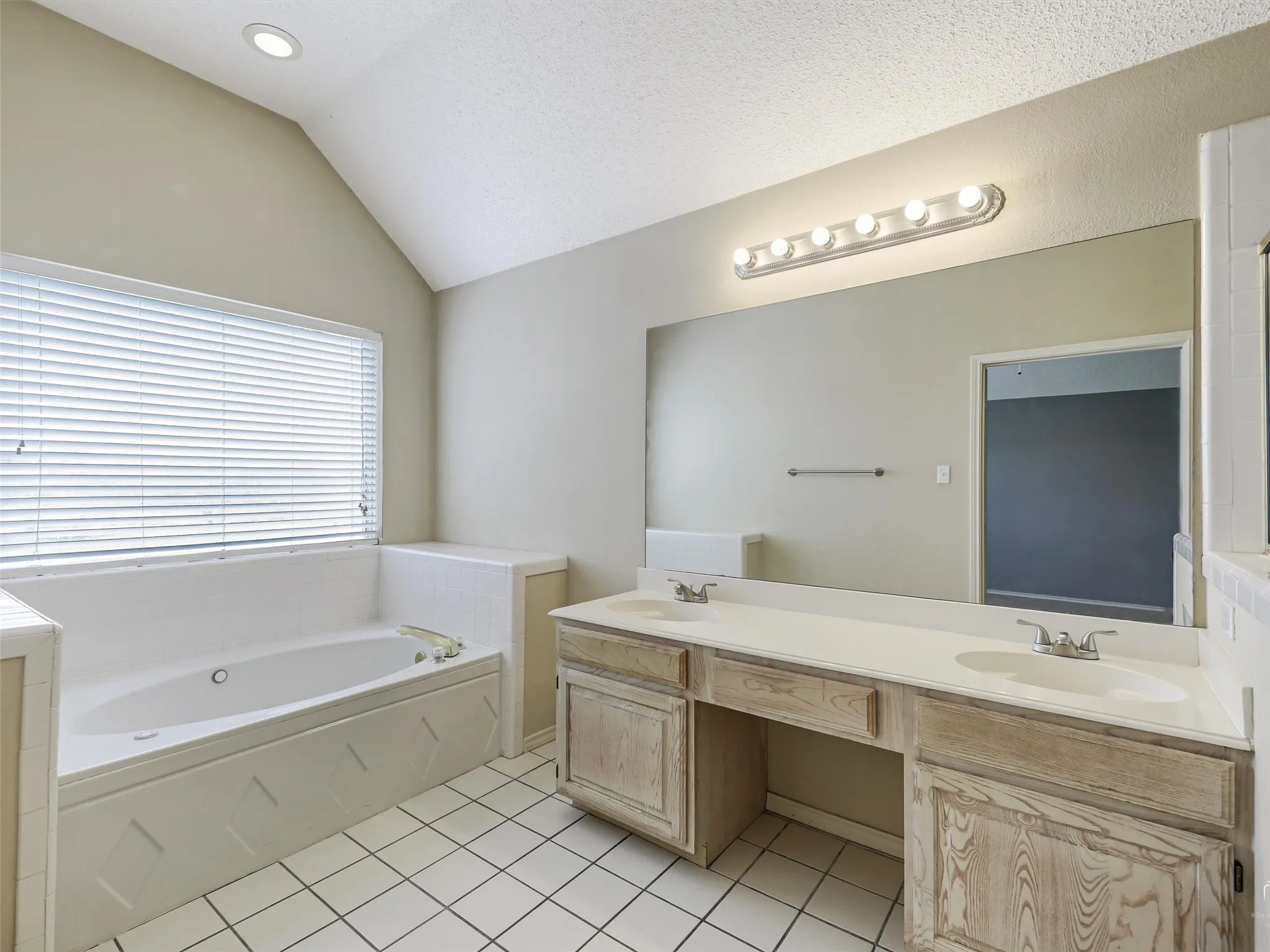 Vaulted ceiling bathroom featuring a dual-sink vanity with a solid-surface countertop, tiled soaking tub surround, recessed lighting, and large wall-mounted mirror