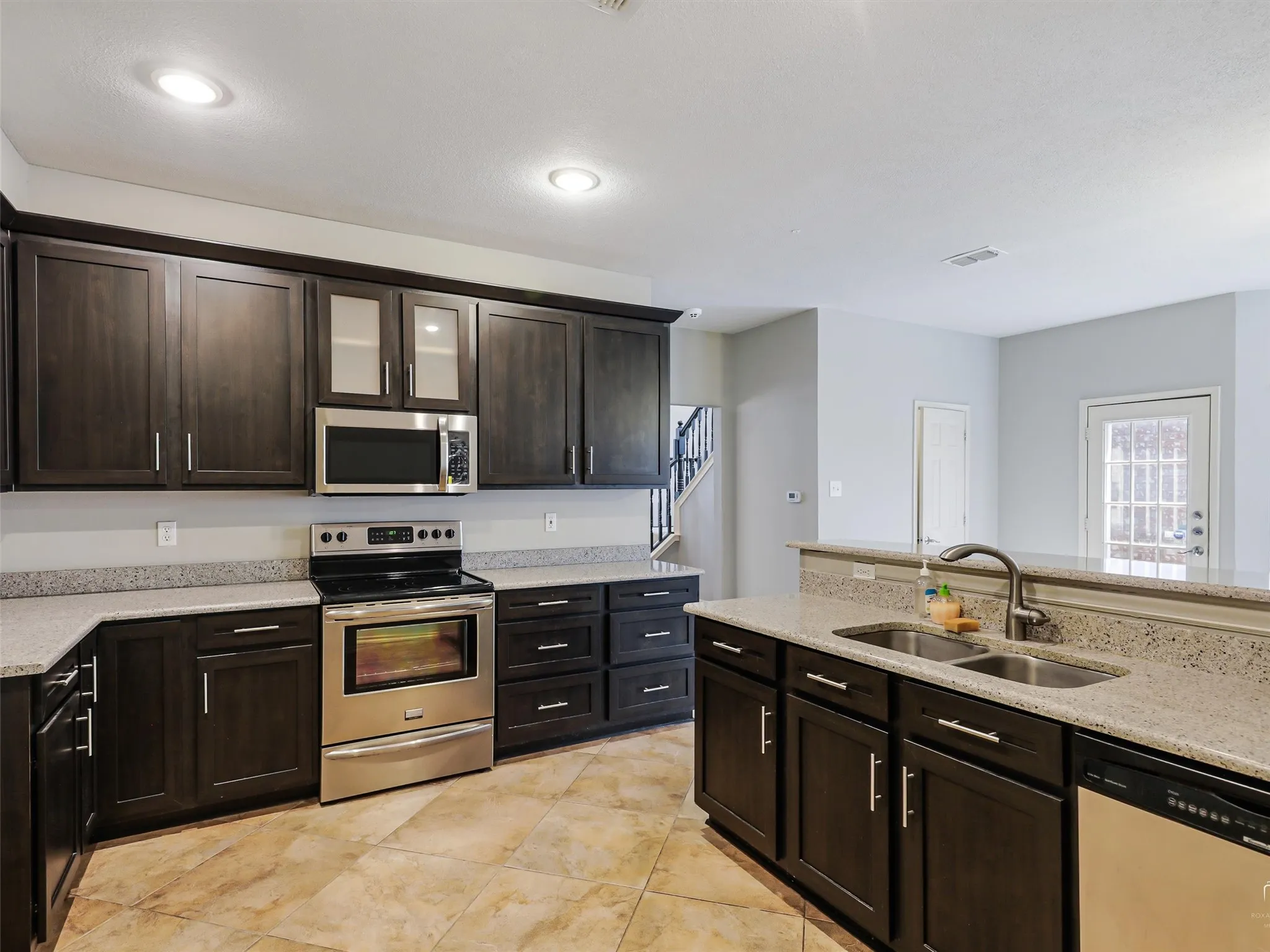 Kitchen featuring dark wood cabinetry, speckled stone countertops, stainless steel appliances, a dual-basin undermount sink with a gooseneck faucet, and tiled flooring
