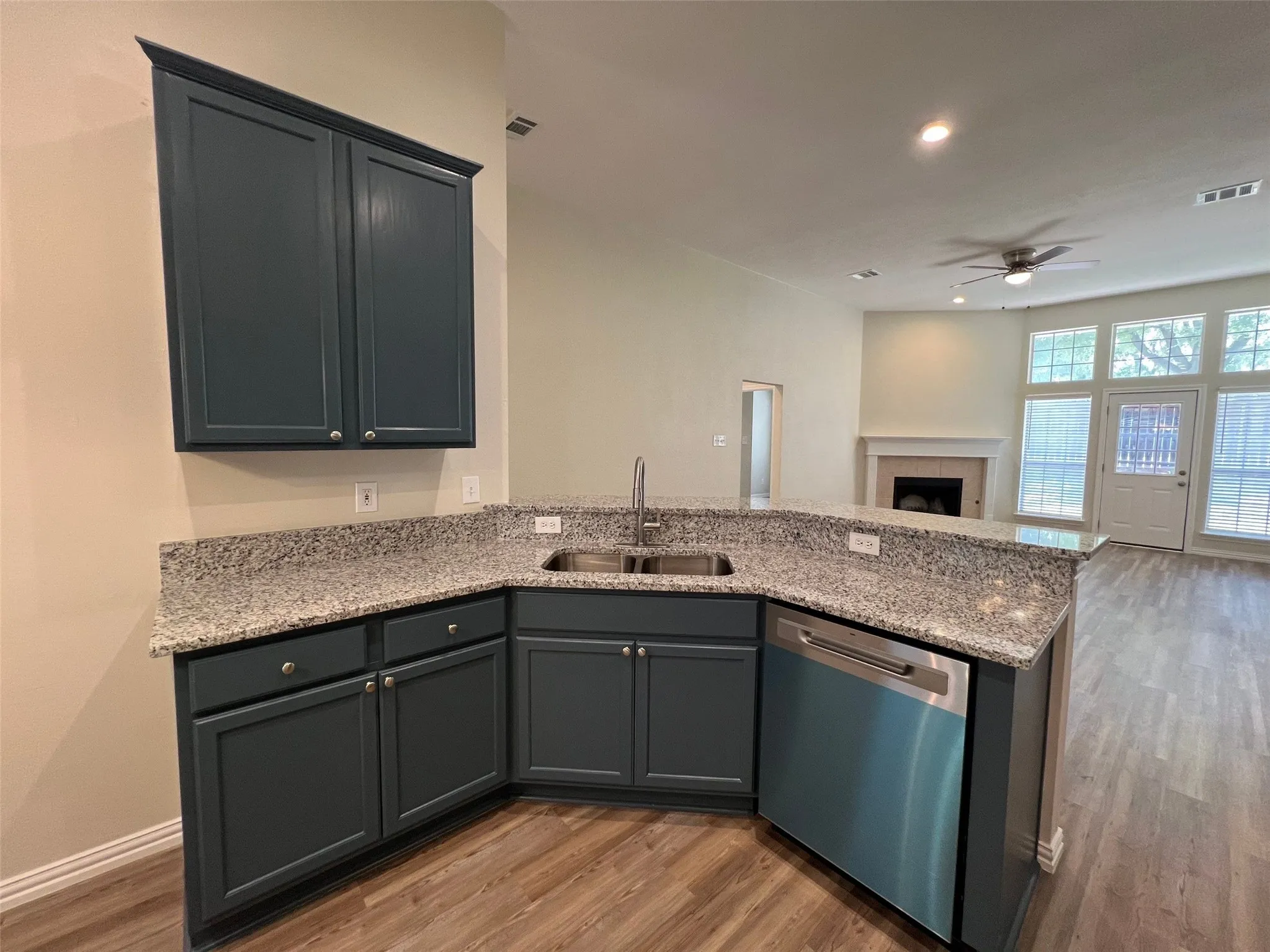 Kitchen area featuring dark-toned cabinetry, stone-surface countertops, dual-basin stainless steel sink, built-in dishwasher, and wood-finish flooring