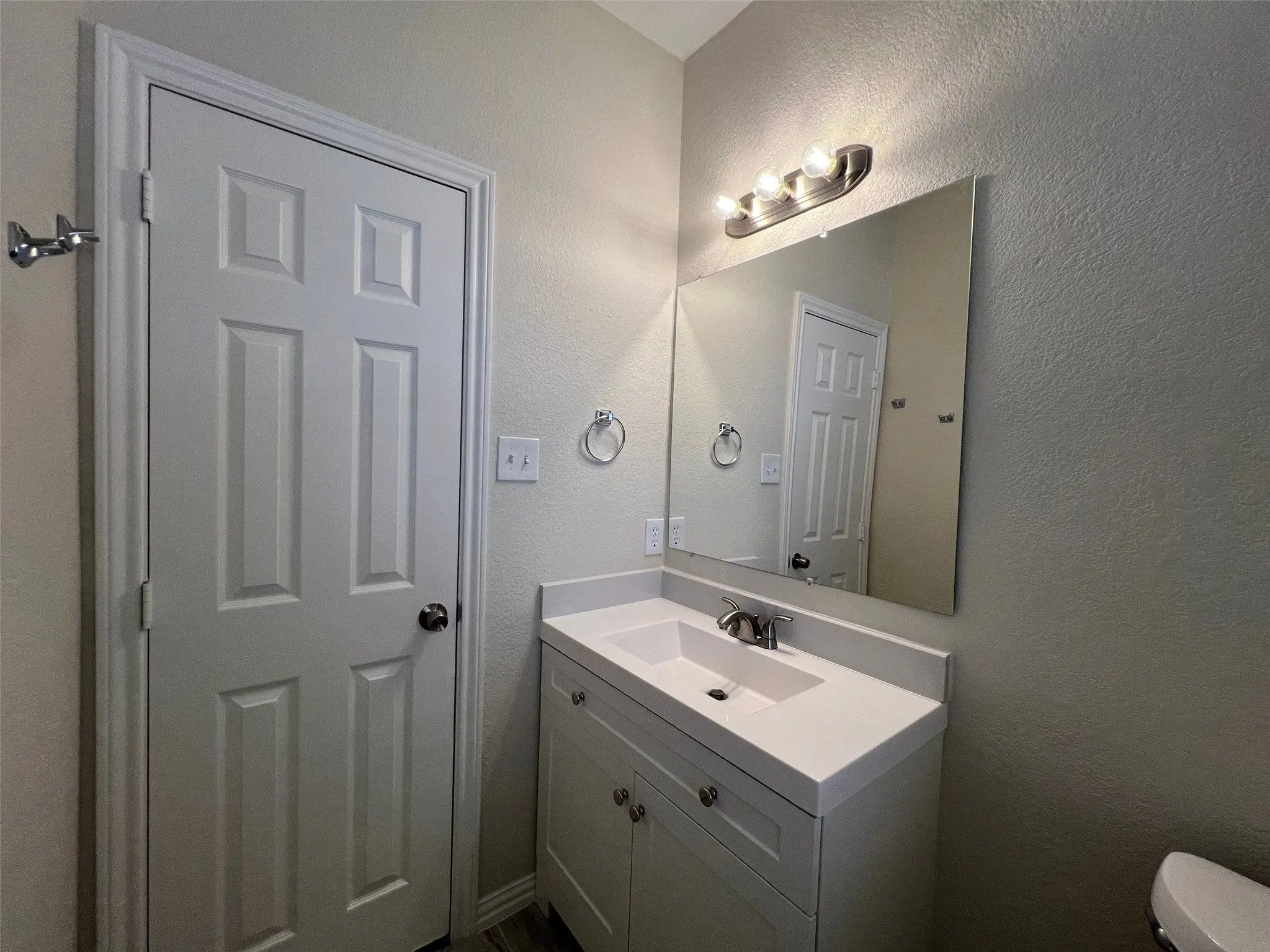 Single-sink vanity featuring a white countertop with integrated basin, shaker-style cabinetry, a large frameless mirror, and a three-light fixture