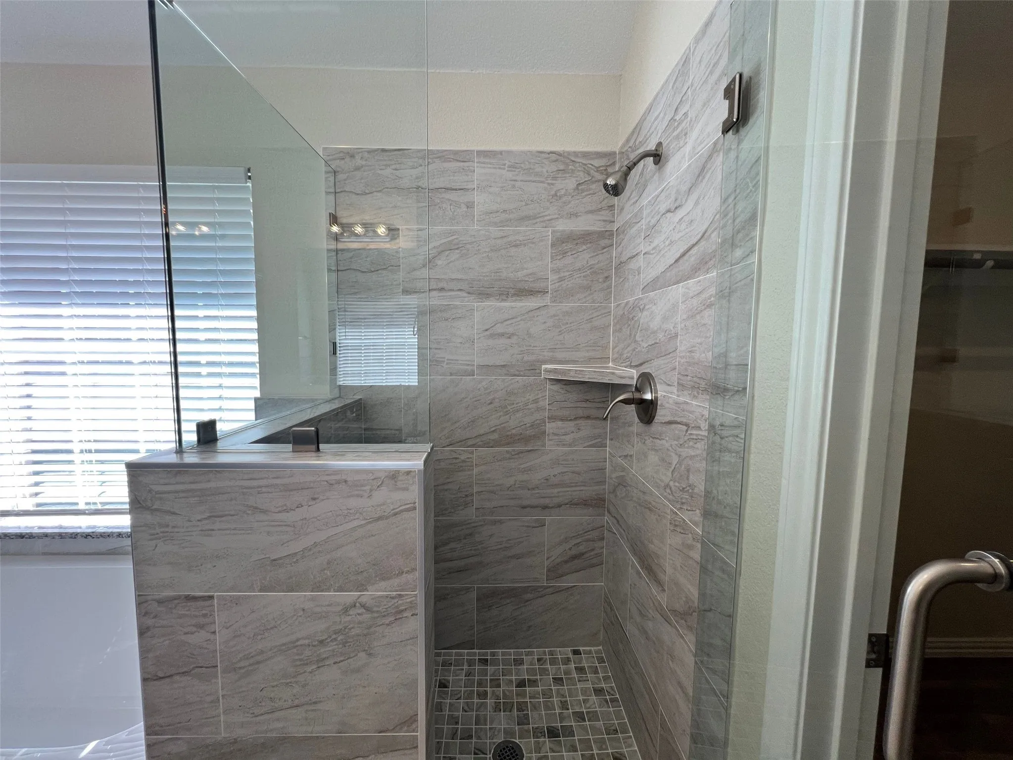Walk-in shower featuring floor-to-ceiling stone-finish tile surround, frameless glass enclosure, built-in corner shelving, mosaic tile flooring, and metallic fixtures