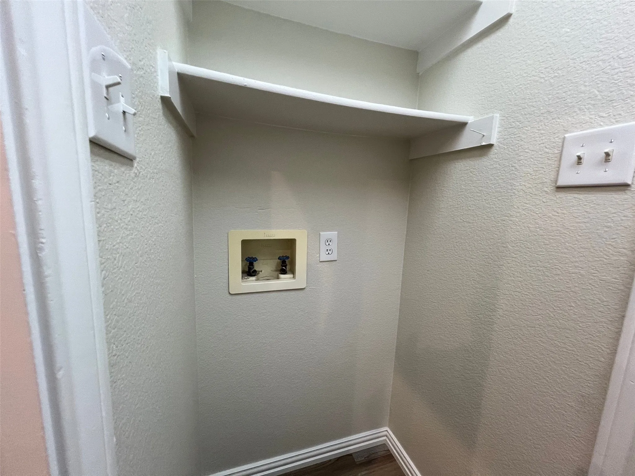 Utility alcove featuring a wall-mounted shelving unit, water supply connection box, and dedicated electrical outlet