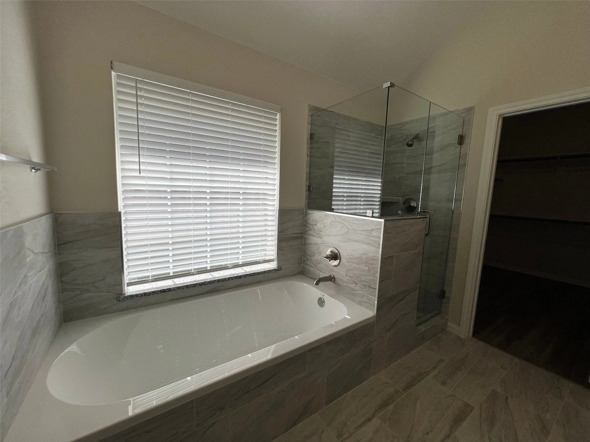Soaking tub with tile surround, glass-enclosed shower, neutral large-format tile flooring, and horizontal window blinds