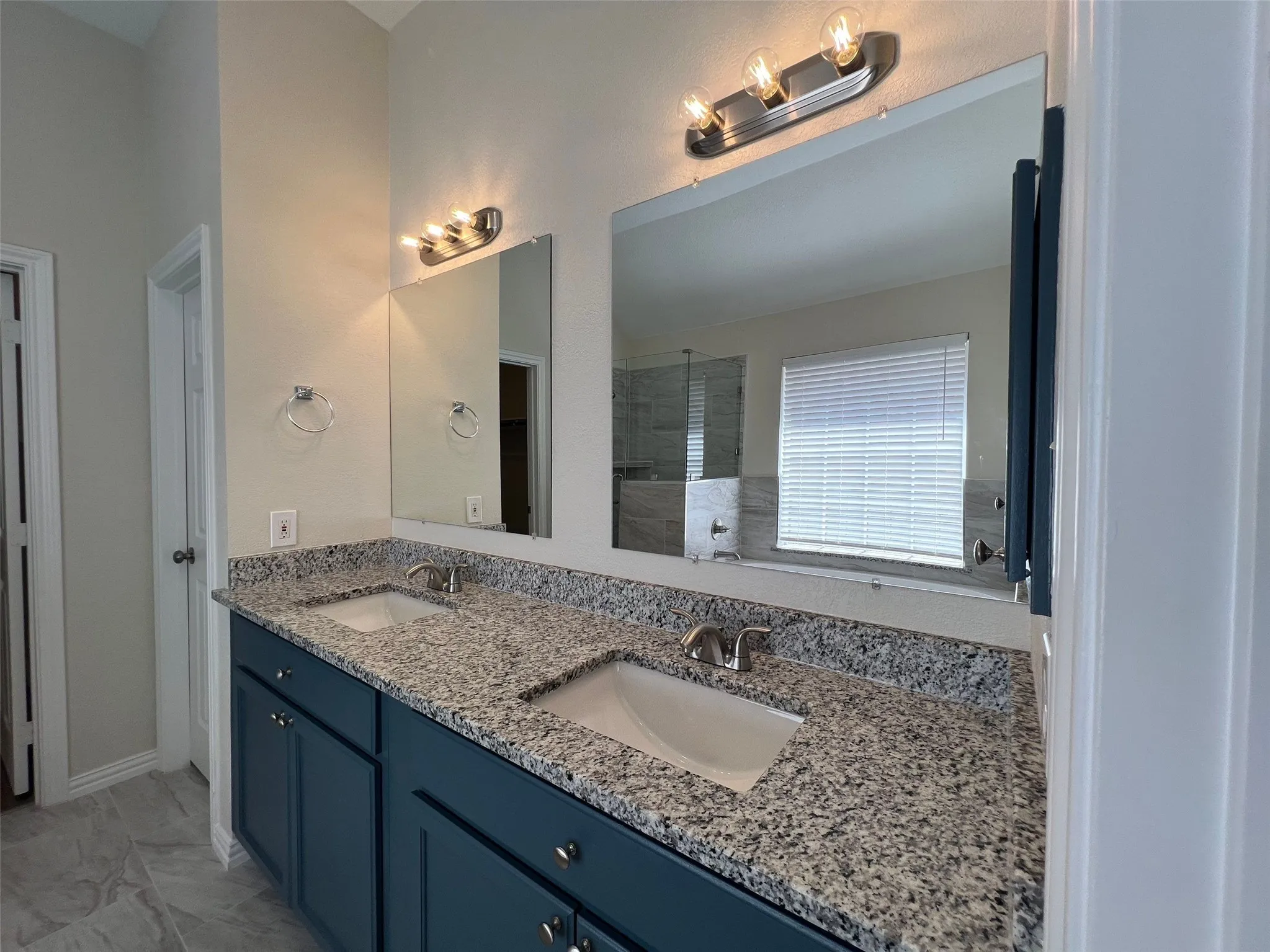 Dual-sink vanity featuring stone-finish countertops, undermount rectangular basins, dark-toned cabinetry, and dual wall-mounted light fixtures with polished metal hardware