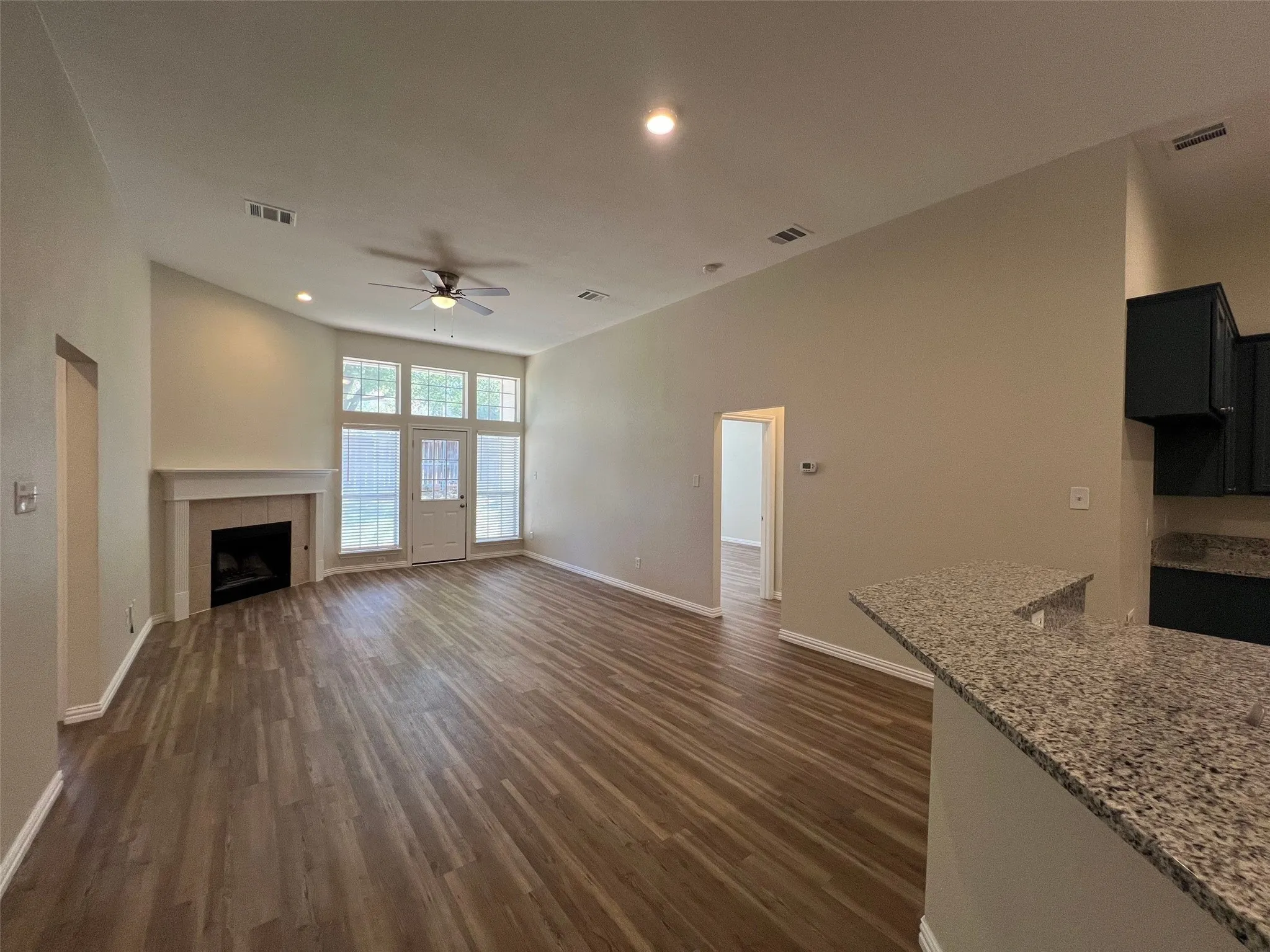 Open-concept living space featuring vaulted ceilings, wood-finish flooring, a fireplace with a decorative mantel, and a kitchen peninsula with stone-finish countertops