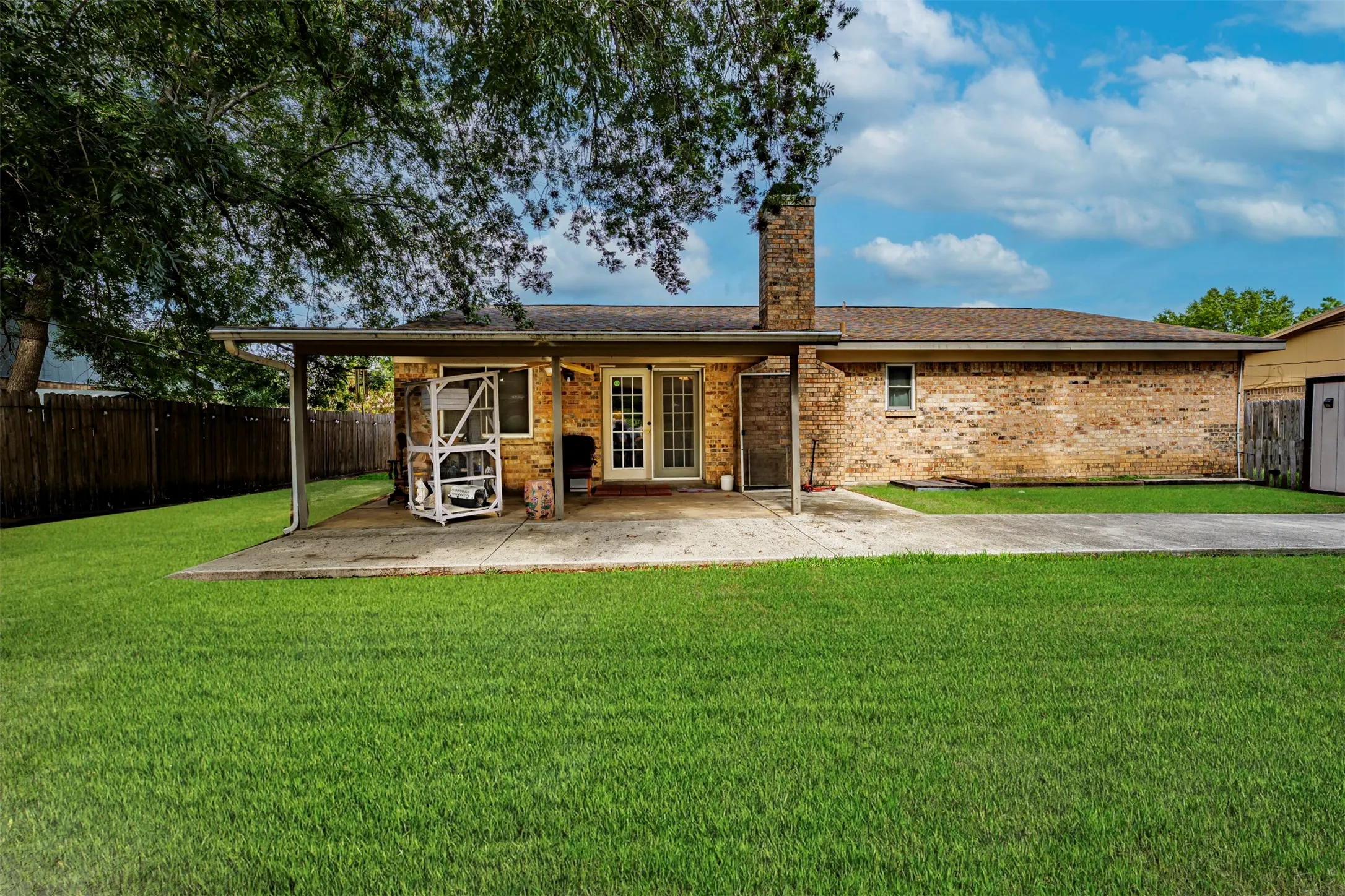 Brick exterior featuring a covered concrete patio, an expansive lawn, a stone chimney, and wood-panel fencing