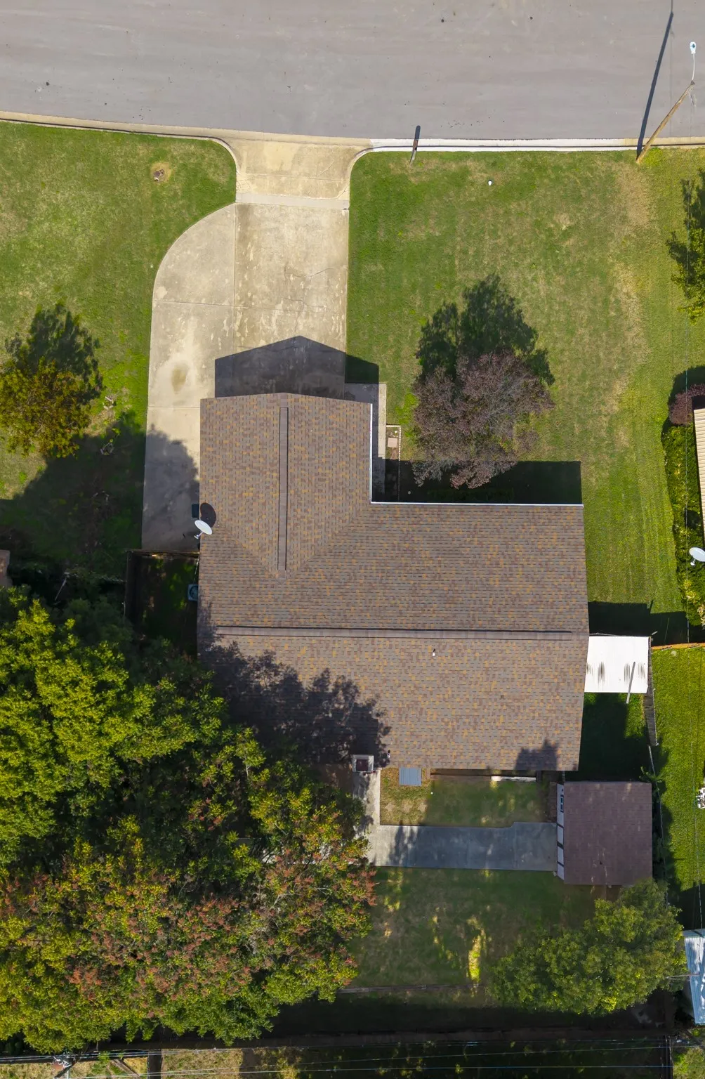Aerial view featuring an asphalt shingle roof, concrete driveway, expansive grass lawn, and mature trees on a corner lot